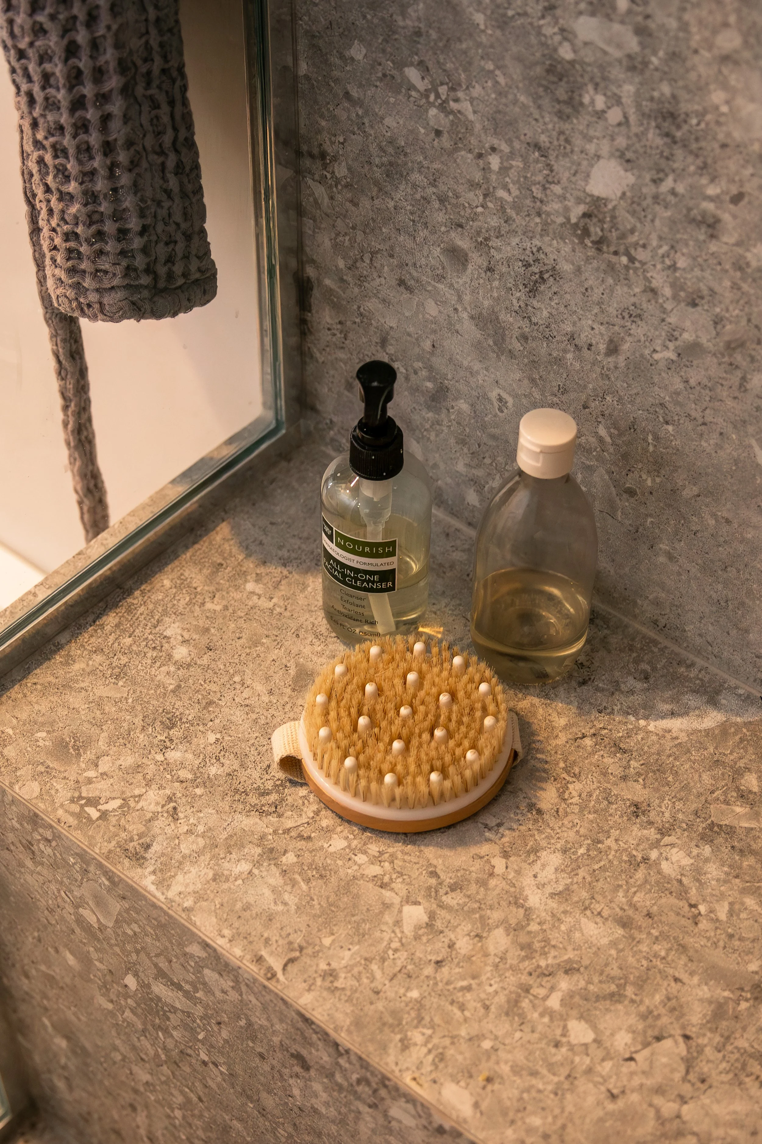 A bathroom countertop with a hand towel hanging on a mirror, a bottle of facial cleanser, a bottle of oil, and a body brush. Bozeman interior design, interior renovation, and interior styling. 
