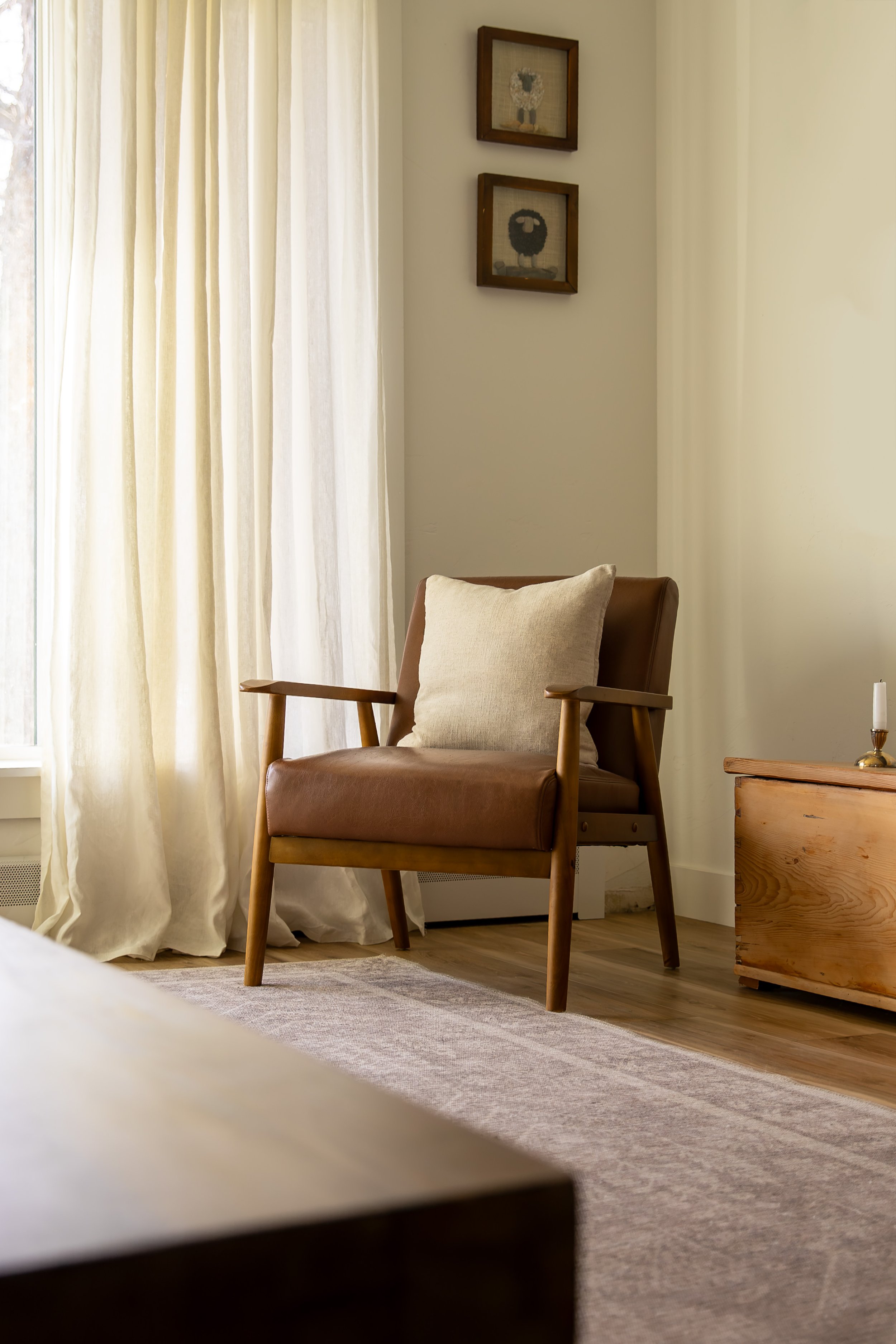 A corner with a leather armchair, beige pillow, wooden side table, and framed artwork on the wall near a window with sheer curtains. Bozeman interior design, interior renovation, and interior styling. 