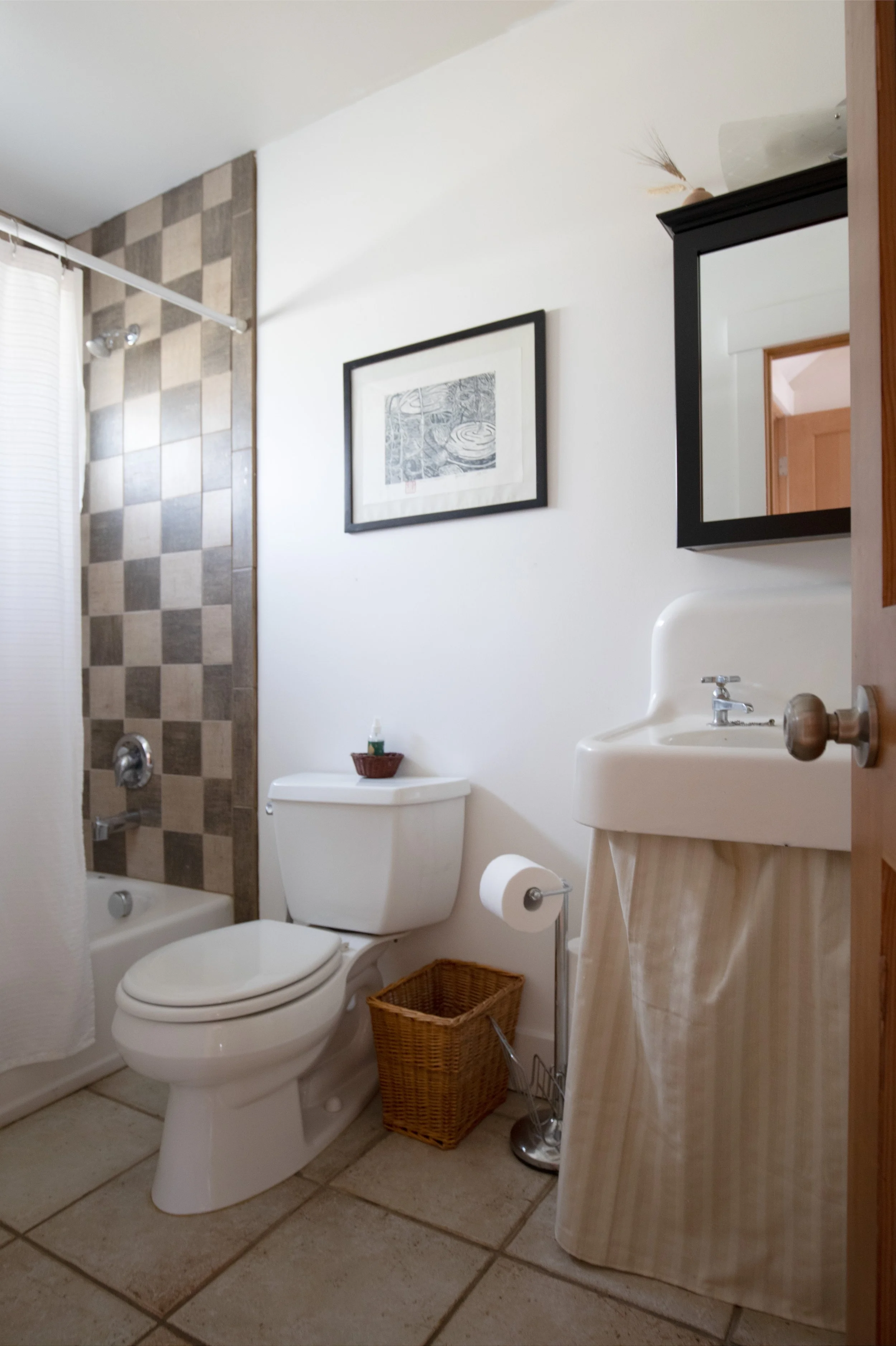 Bathroom with a shower, toilet, small sink with striped curtain, framed art, and wicker basket. bozeman montana two dot interiors