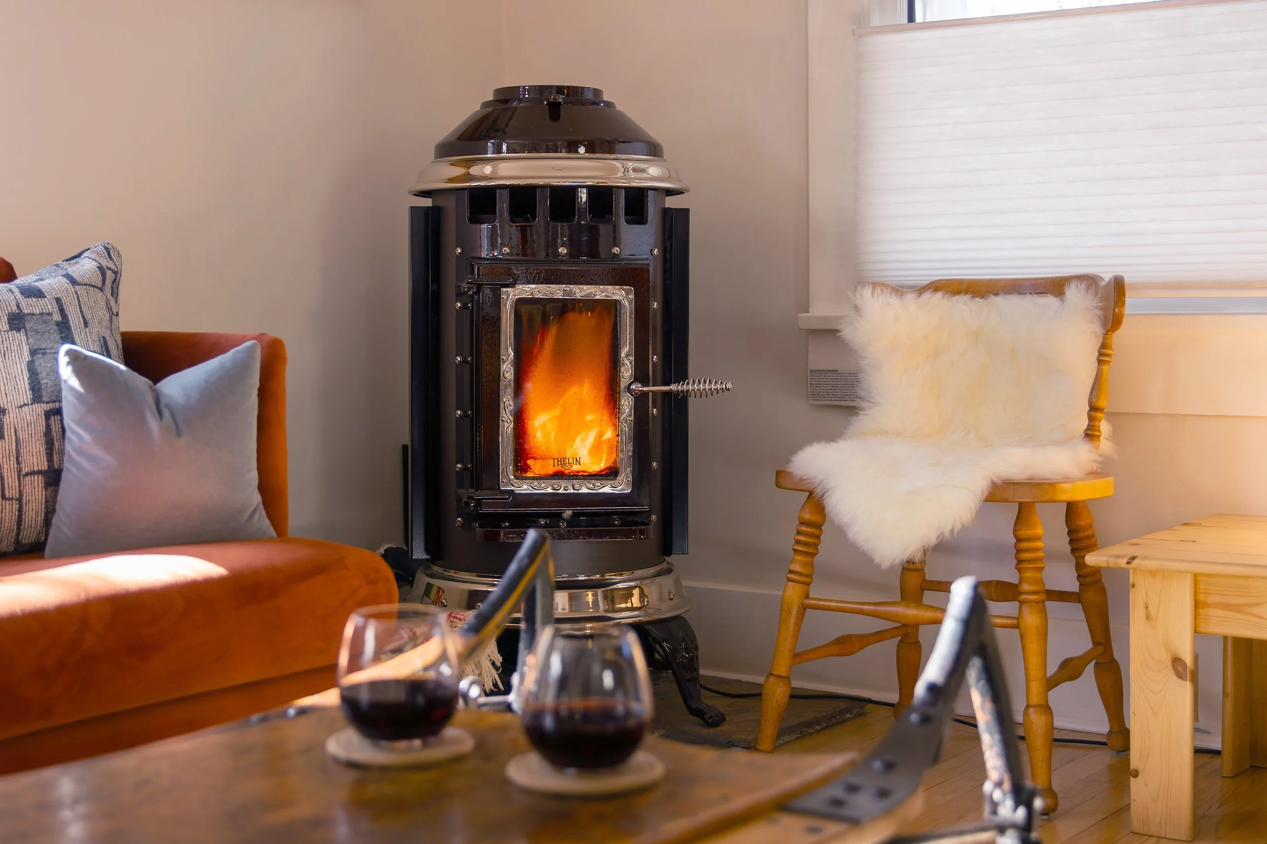 A cozy living room corner with a wood-burning stove emitting flames, an orange couch with throw pillows, a wooden chair with a white furry throw, and a window with blinds. refresh renovation remodel interior designer bozeman montana two dot interiors