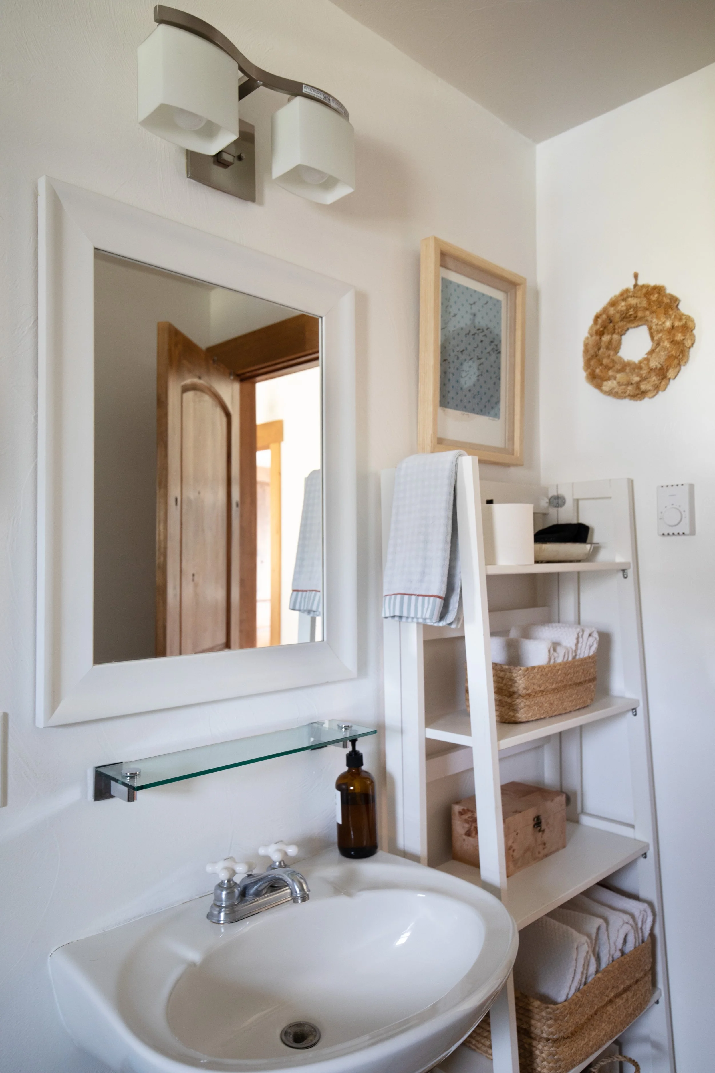 A bathroom with a white sink, a mirror above the sink, a glass shelf below the mirror, a towel hanging on a ladder, a wooden shelf unit with towels and baskets, a framed artwork, a decorative wreath, and a ceiling light fixture.