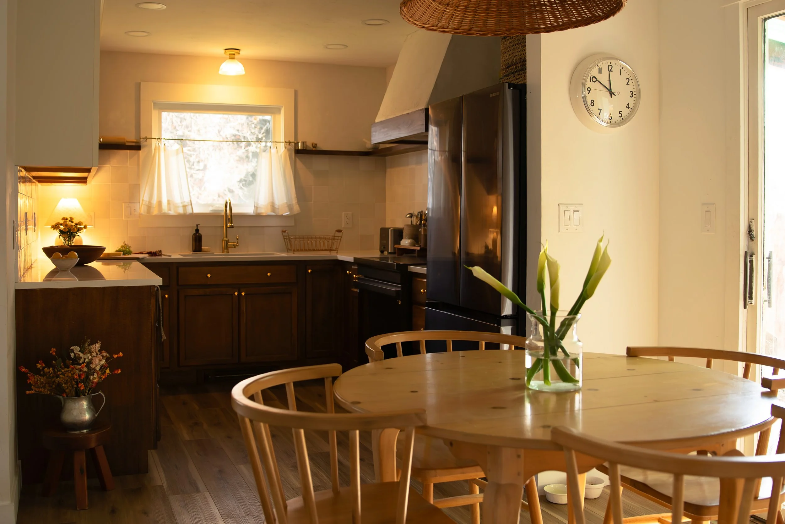 A cozy kitchen with a wooden dining table with chairs and a vase of white lilies, dark wood cabinets, and a window with white curtains letting in natural light. Bozeman interior design, interior renovation, and interior styling.