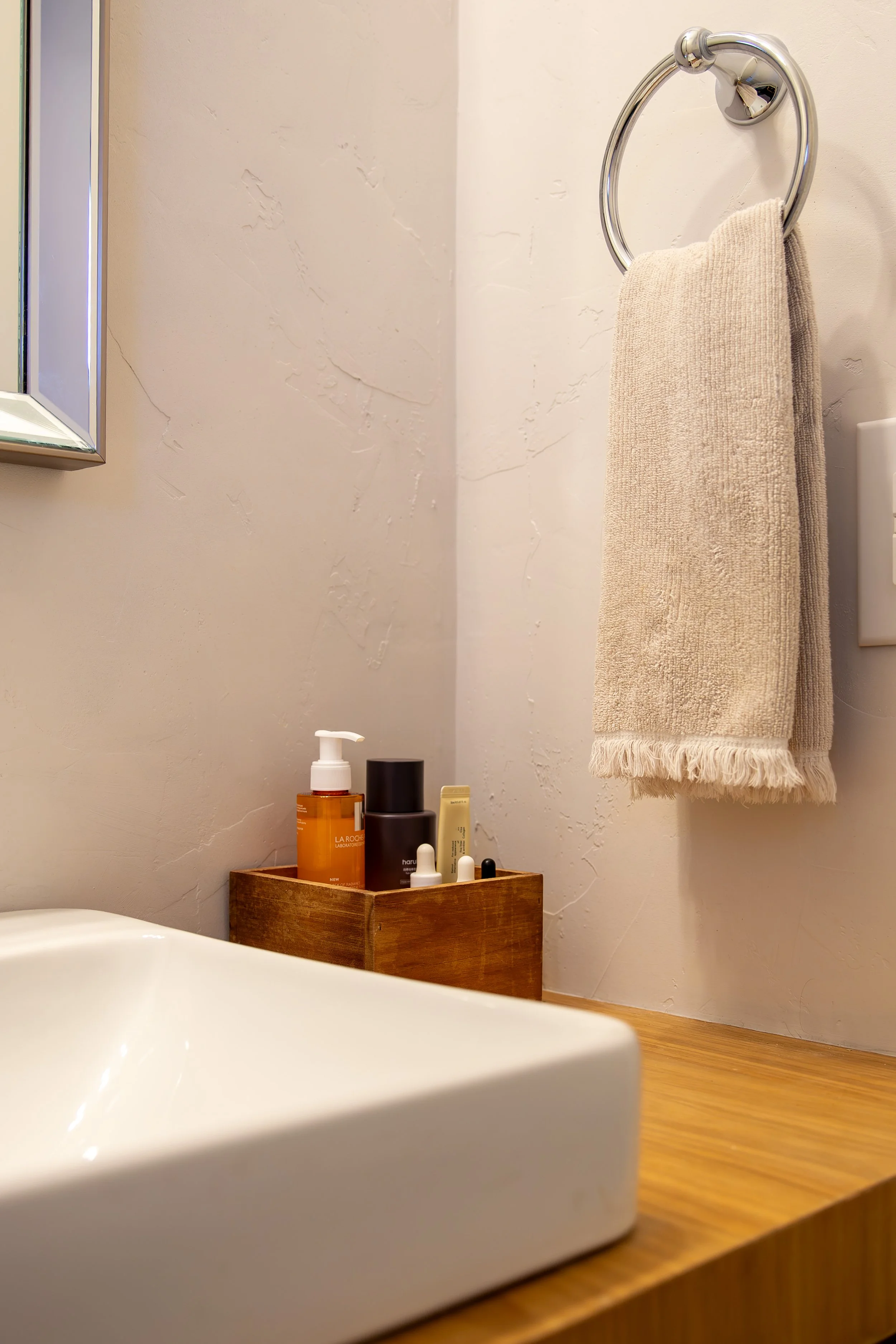 Close-up of a modern bathroom with a white oak countertop, a beige towel hanging on a round chrome towel ring, and a wooden tray containing various skincare products. Bozeman interior design, interior renovation, and interior styling.
