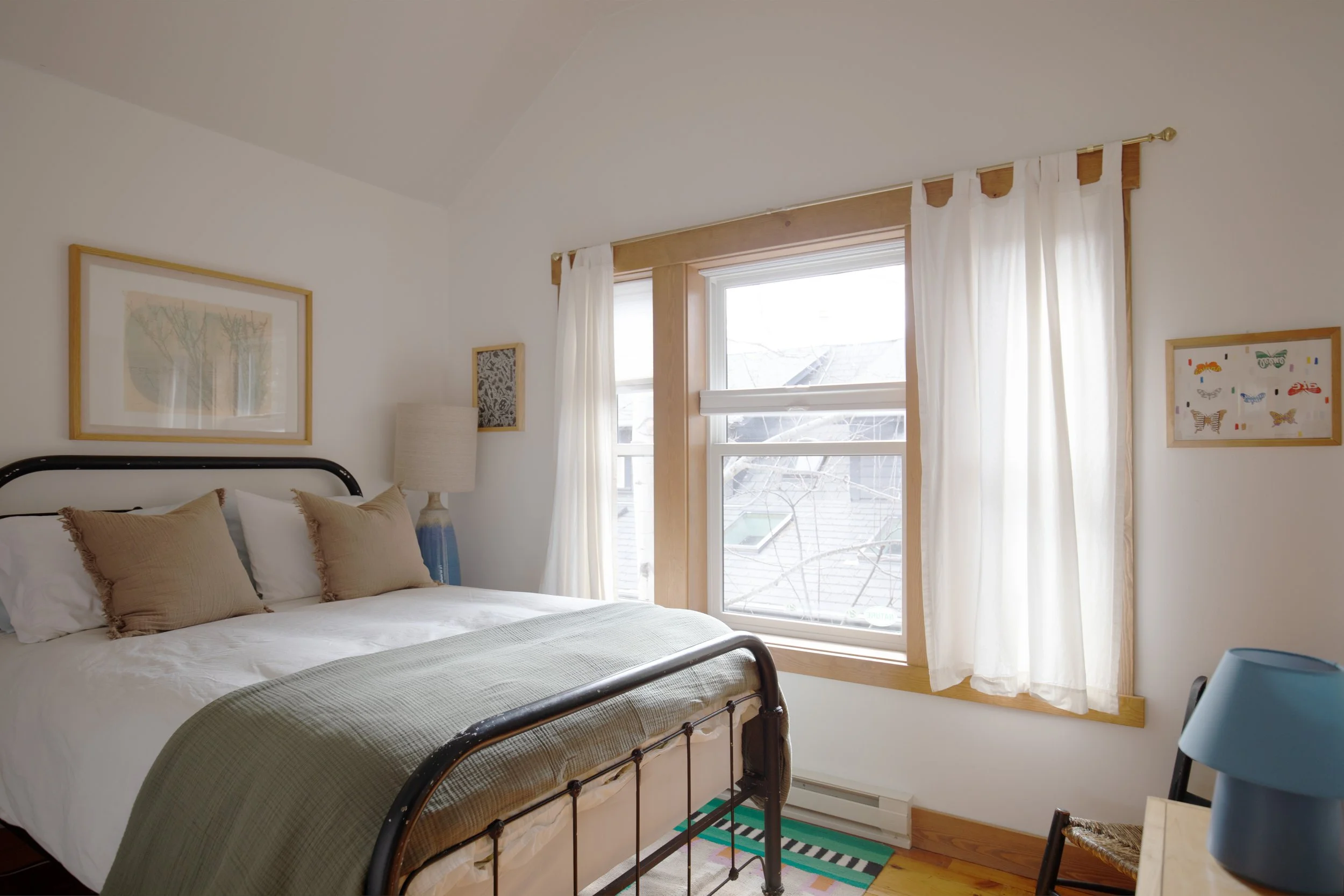 A bedroom with a large bed, beige pillows, and a green blanket, next to a window with white curtains, a lamp, and wall art. STR refresh interior design bozeman montana two dot interiors