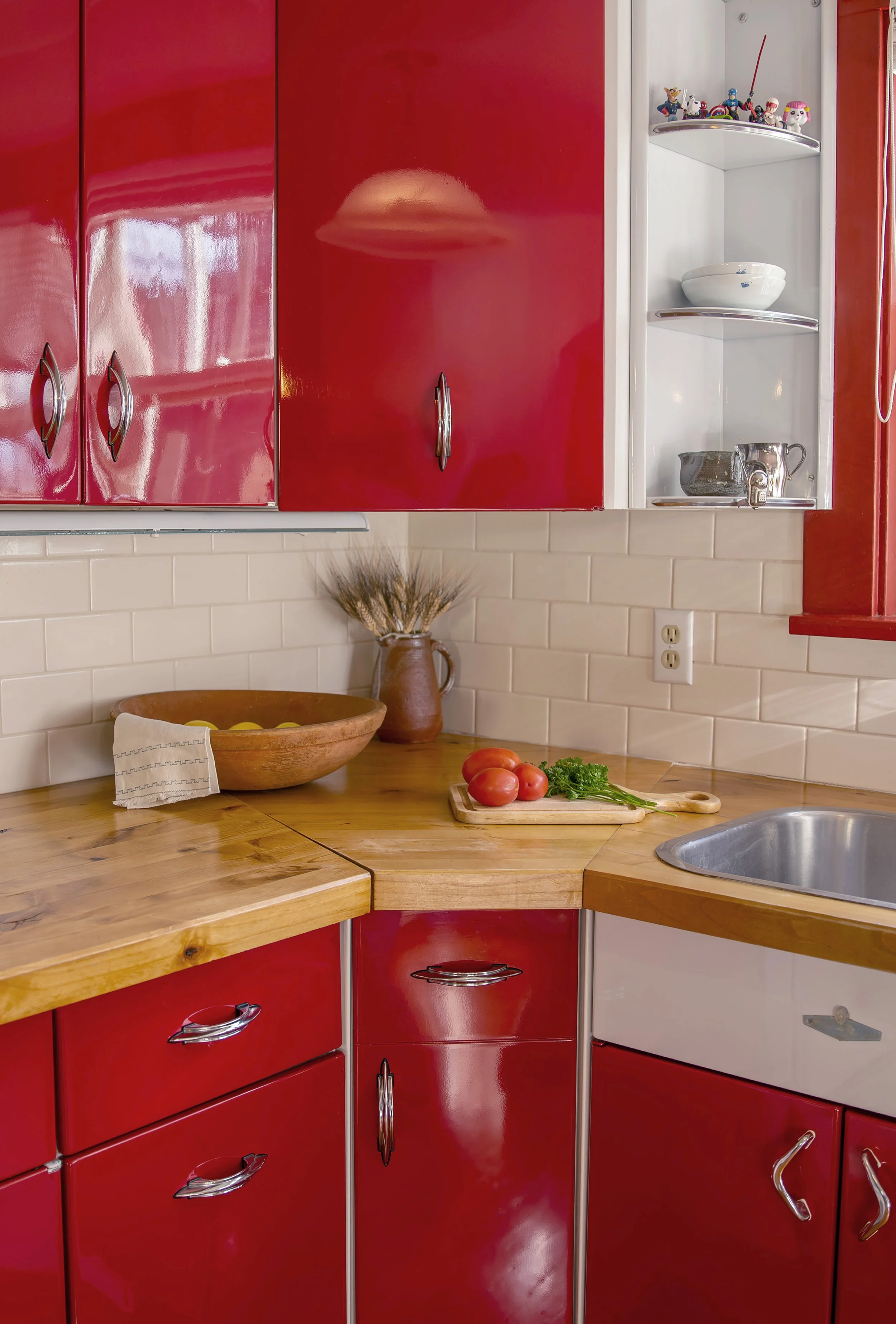 A kitchen with red cabinets, wooden countertop, white brick backsplash, and a small open shelving unit with toys, dishes, and a pitcher. The countertop has tomatoes, parsley, a wooden bowl with lemons, a ceramic pitcher, and a vase with wheat stalks.