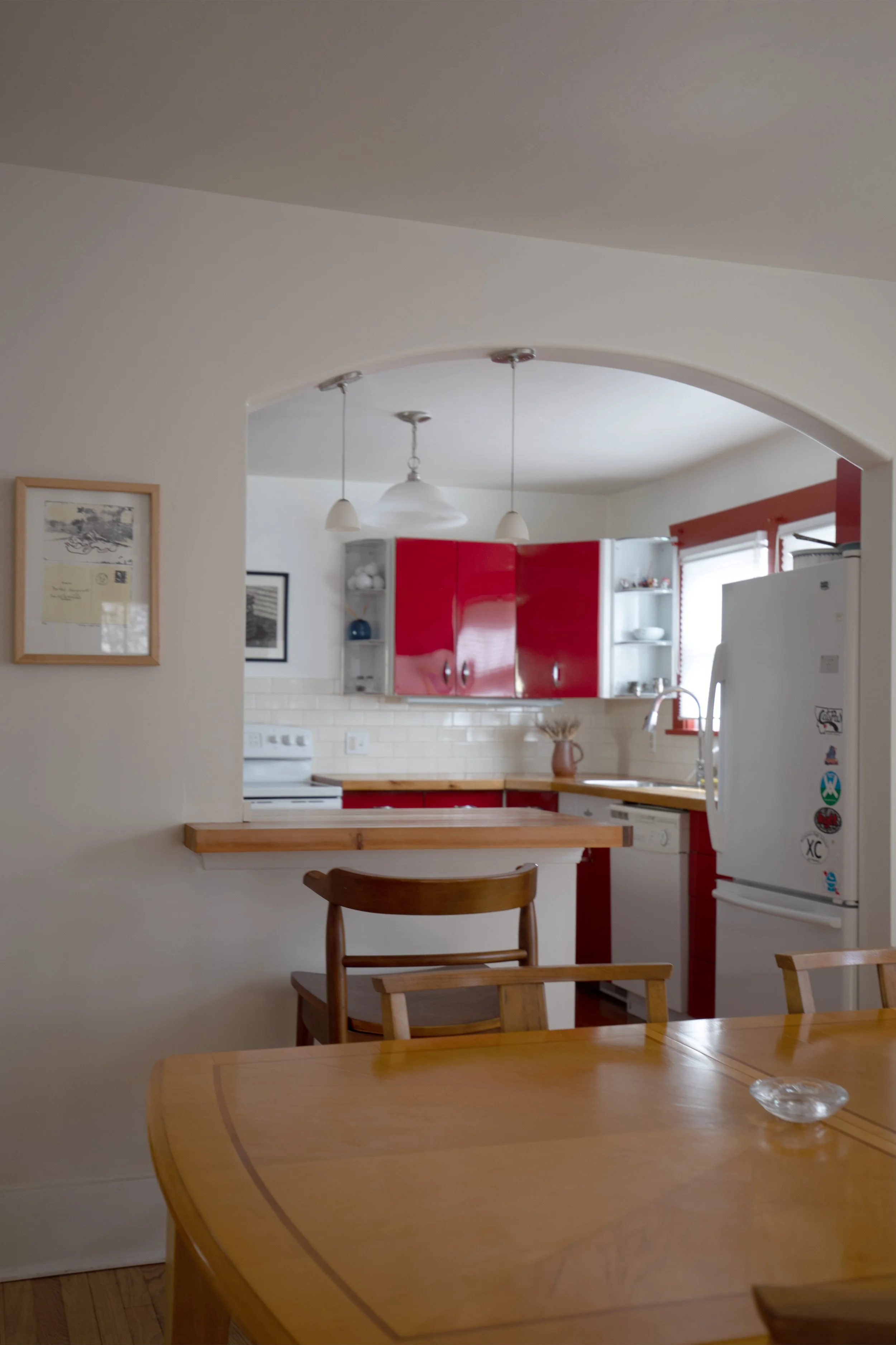 View of a kitchen with red cabinets and white appliances, seen through an arched doorway from a dining area with a wooden table and chairs.