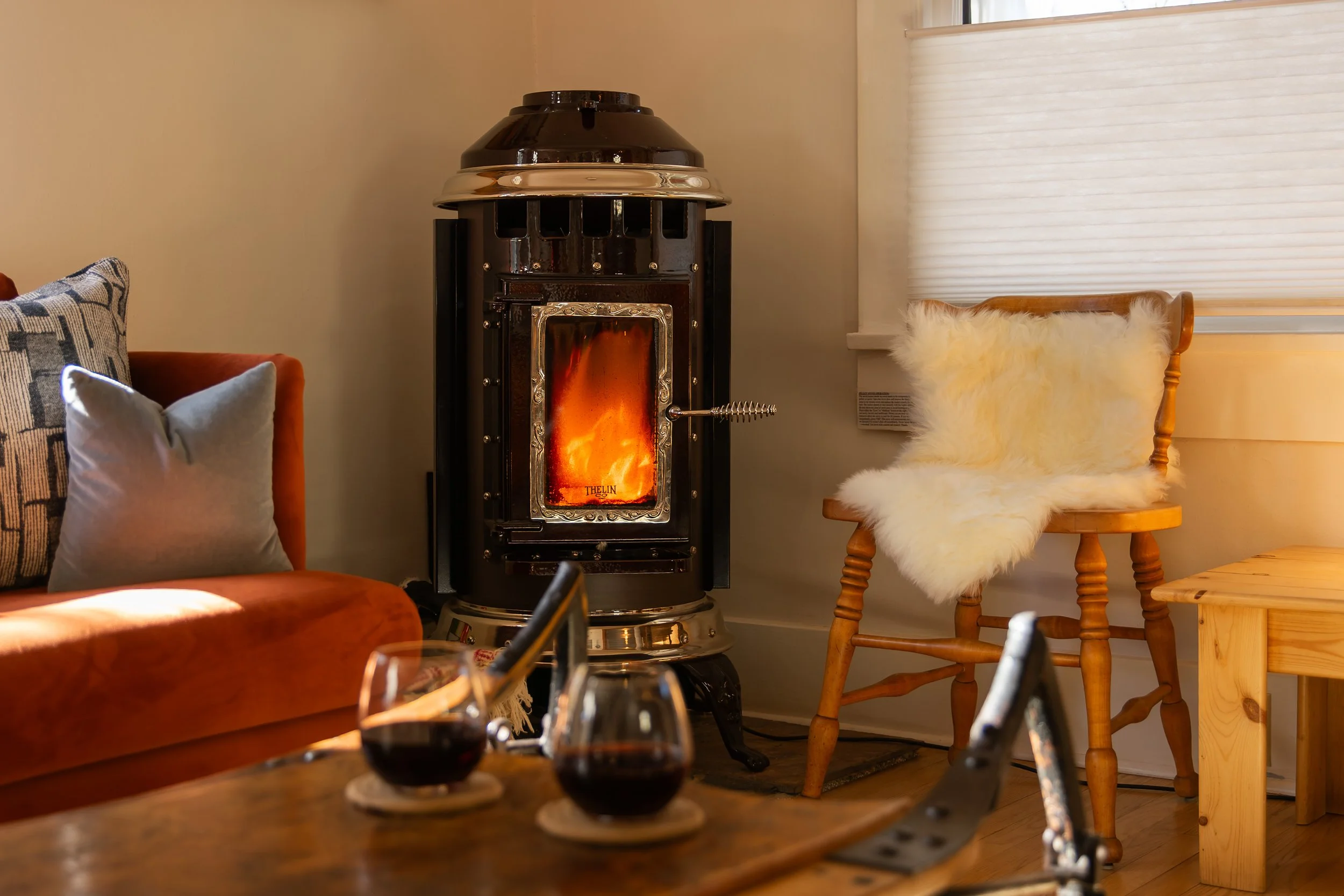 A cozy living room scene with a black wood stove burning fire, a wooden chair with a fluffy cream-colored throw, a rust-colored sofa with pillows, a partial view of a wooden table with two glasses of dark red wine, and a window with closed blinds.