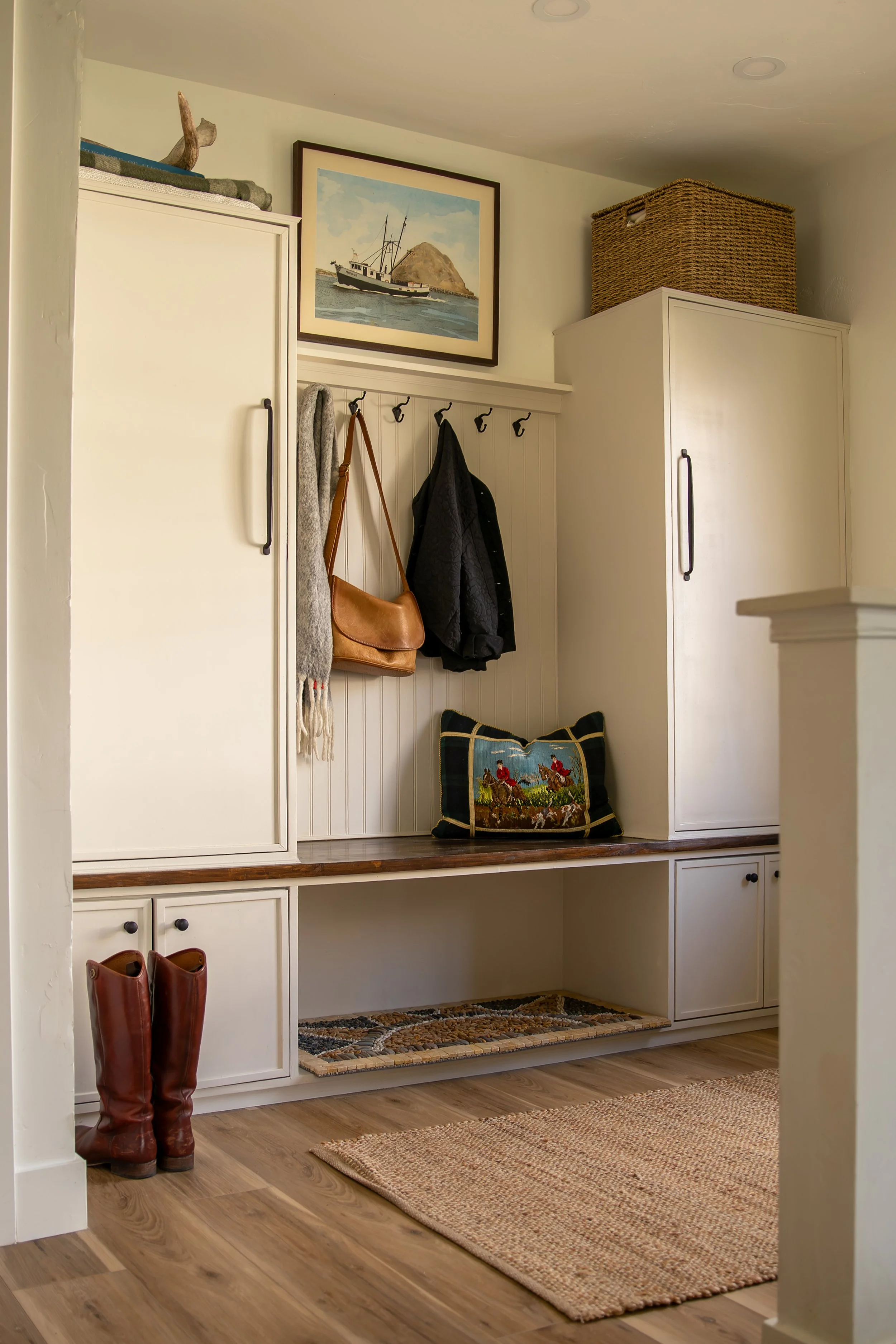 Entryway with white built-in cabinets, coat hooks with jackets, scarf, and purse, decorative pillow, framed nautical painting, wicker basket on top, and a woven rug. Bozeman interior design, interior renovation, and interior styling. 
