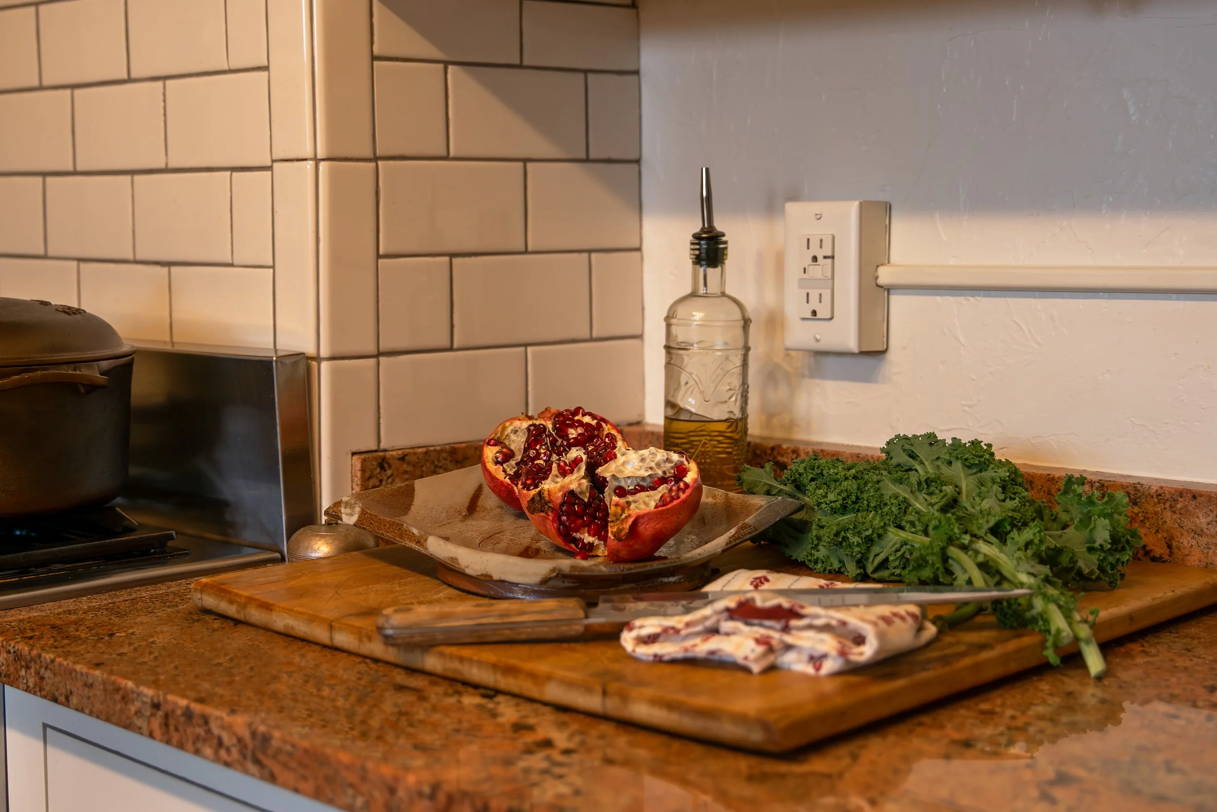 A kitchen countertop with a halved pomegranate, chopped kale, a glass oil dispenser, and a knife on a wooden cutting board. Bozeman interior design, interior renovation, and interior styling. 