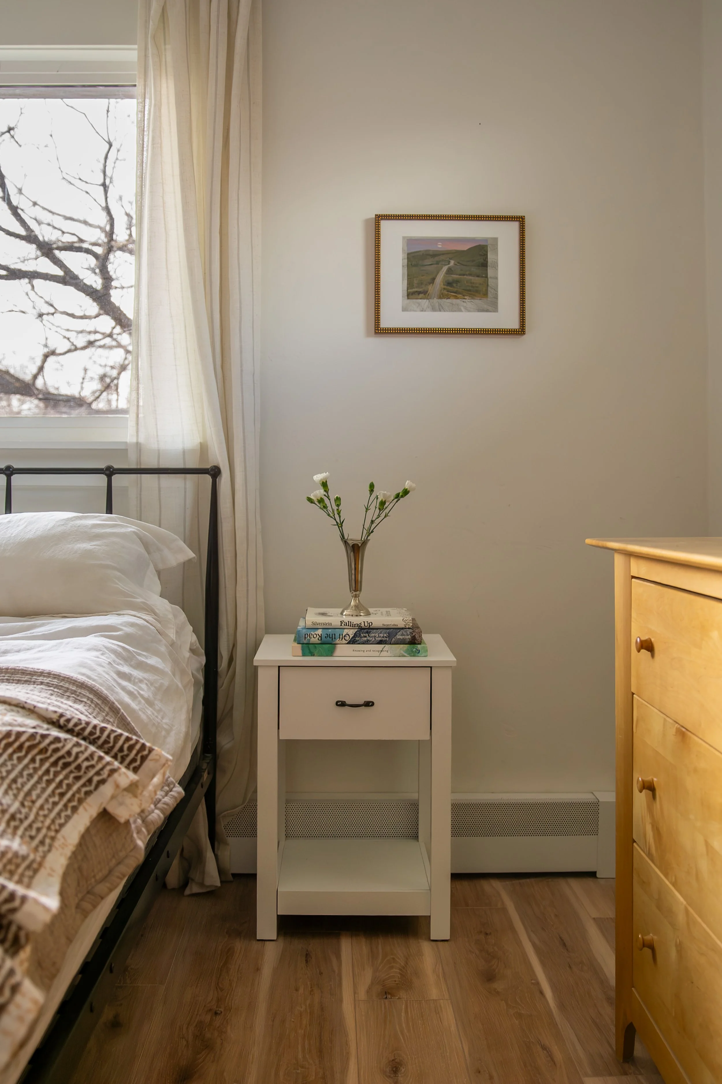 A bedroom corner with a bed, a small white nightstand with a vase and books, a framed picture on the wall, a window with curtains, and a wooden dresser on a wooden floor. Bozeman interior design, interior renovation, and interior styling. 