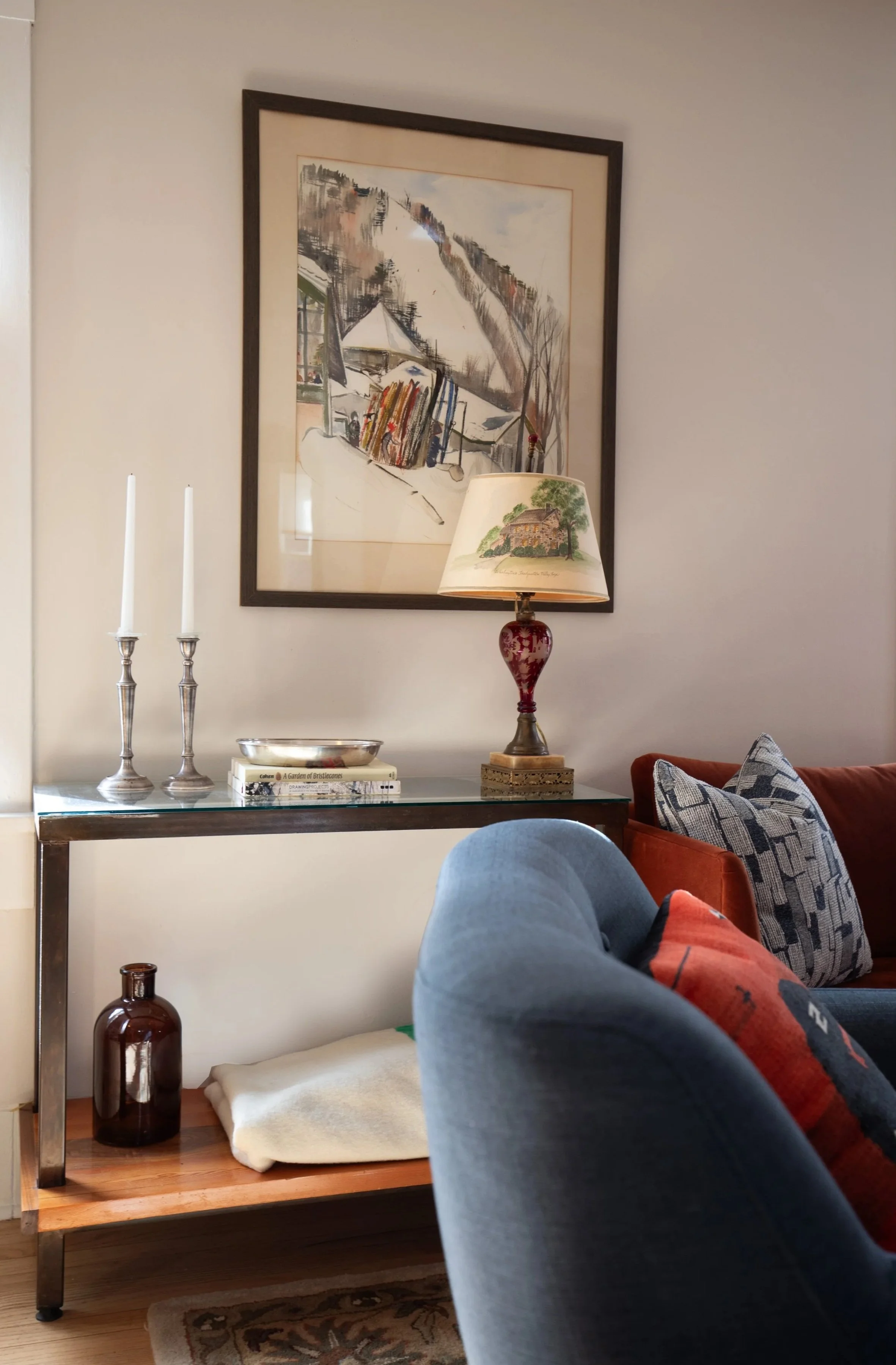 Living room corner with a glass console table holding candles, books, a decorative lamp with painted shade, and a framed artwork. There are patterned pillows on a red armchair and a blue sofa nearby. antiques two dot interiors