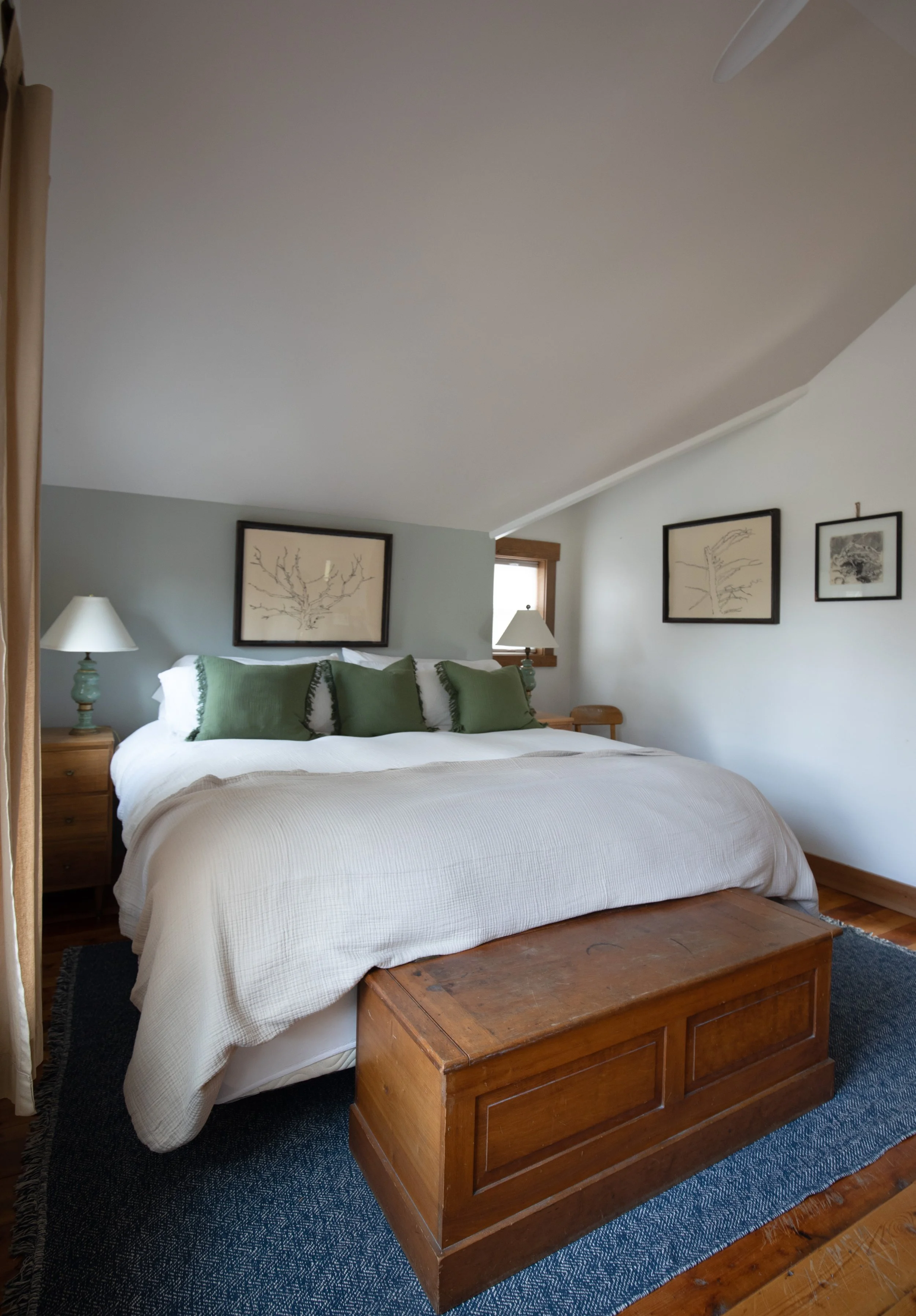 A cozy bedroom with a large bed, white bedding, four green pillows, wooden nightstands with lamps, framed artwork on the walls, a small window, and a wooden chest at the foot of the bed. bozeman montana two dot interiors