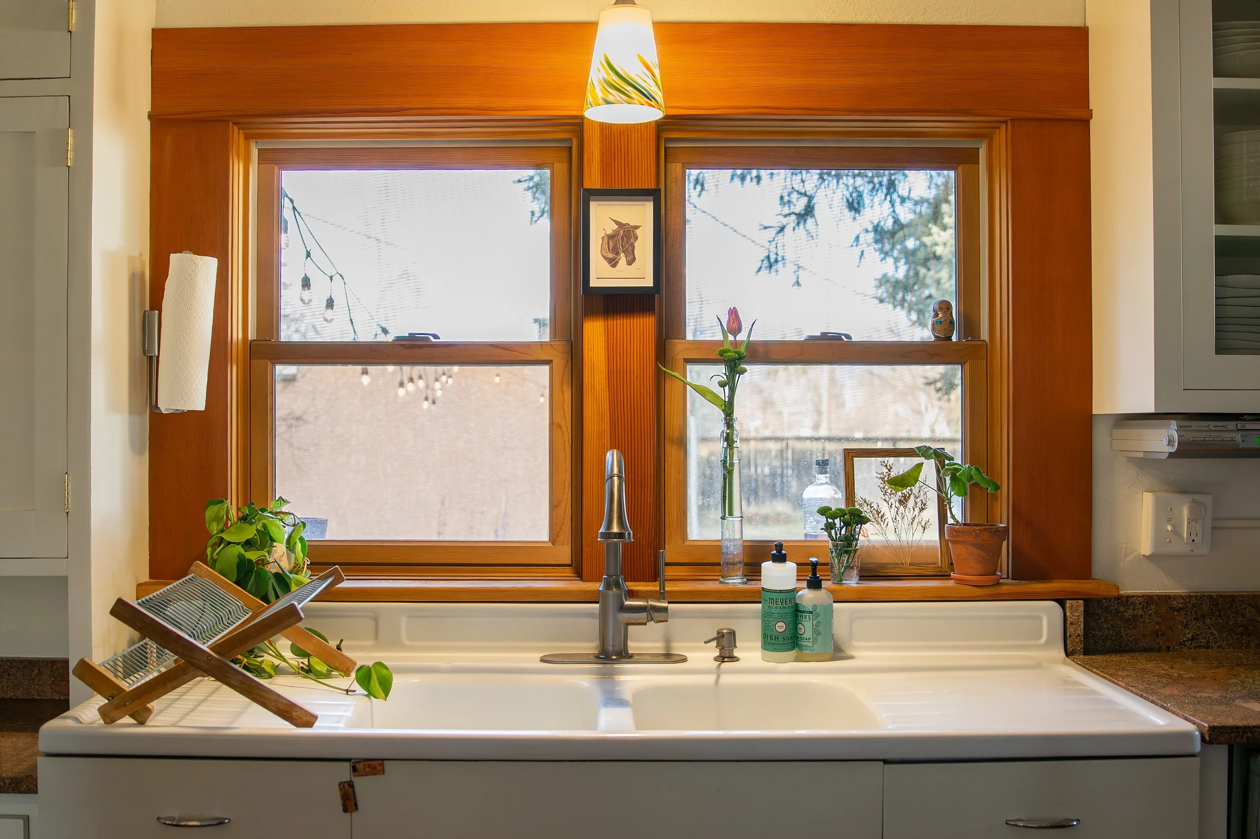 Kitchen sink with a window above, decorated with plants, framed artwork, and bottles, with a wooden window frame and natural light. Bozeman interior design, interior renovation, and interior styling. 