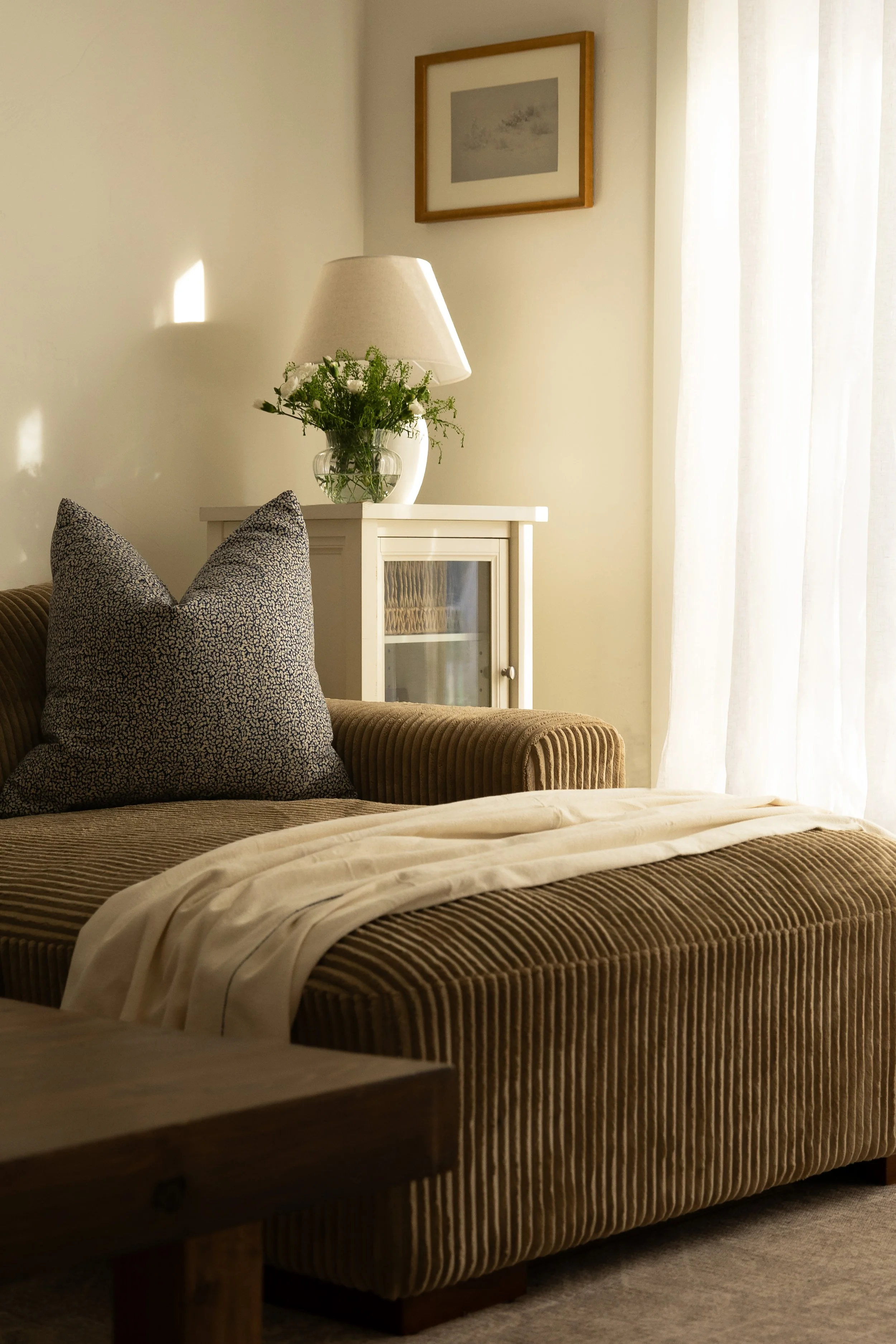 A living room with a sofa, a white cabinet with glass door, a table lamp with a white shade, and a vase with green plants on top, with sheer white curtains. Bozeman interior design, interior renovation, and interior styling. 