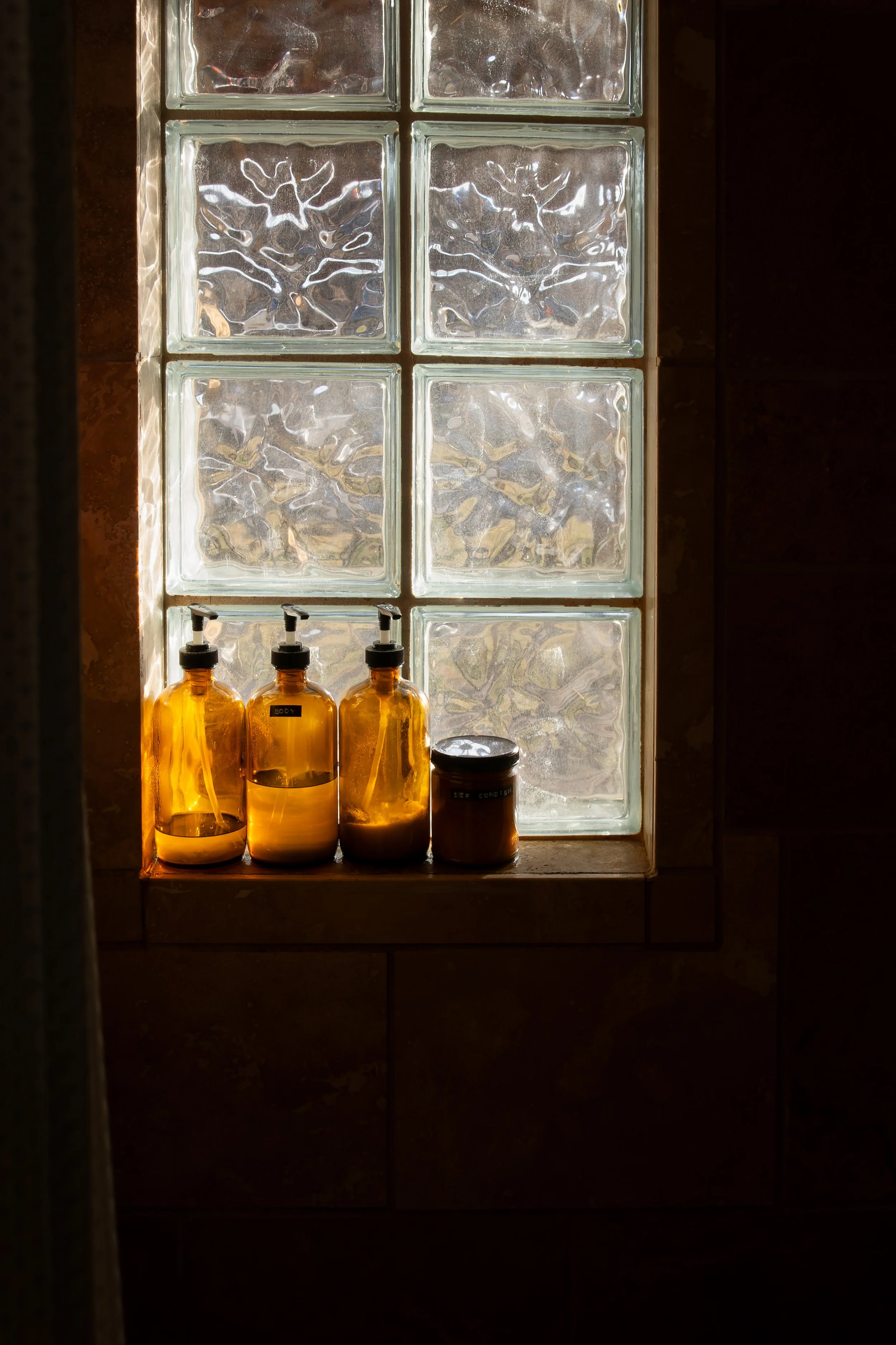 A window made of glass blocks with sunlight shining through, revealing four amber glass bottles with pump dispensers and a small jar on the windowsill. Bozeman interior design, interior renovation, and interior styling. 