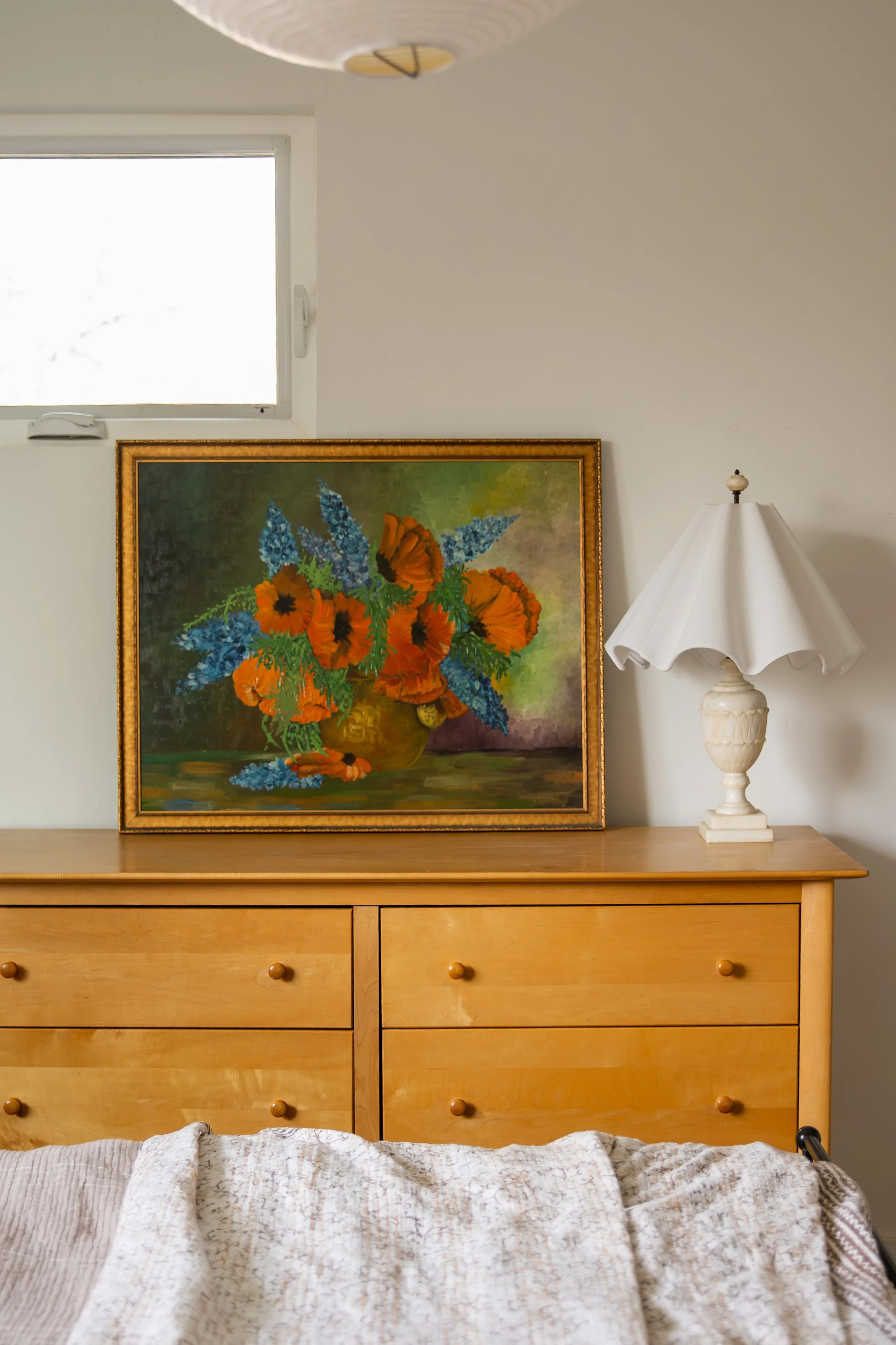 A framed painting of a colorful flower bouquet on a wooden dresser, with a white table lamp to the right and a window in the background. Bozeman interior design, interior renovation, and interior styling. 