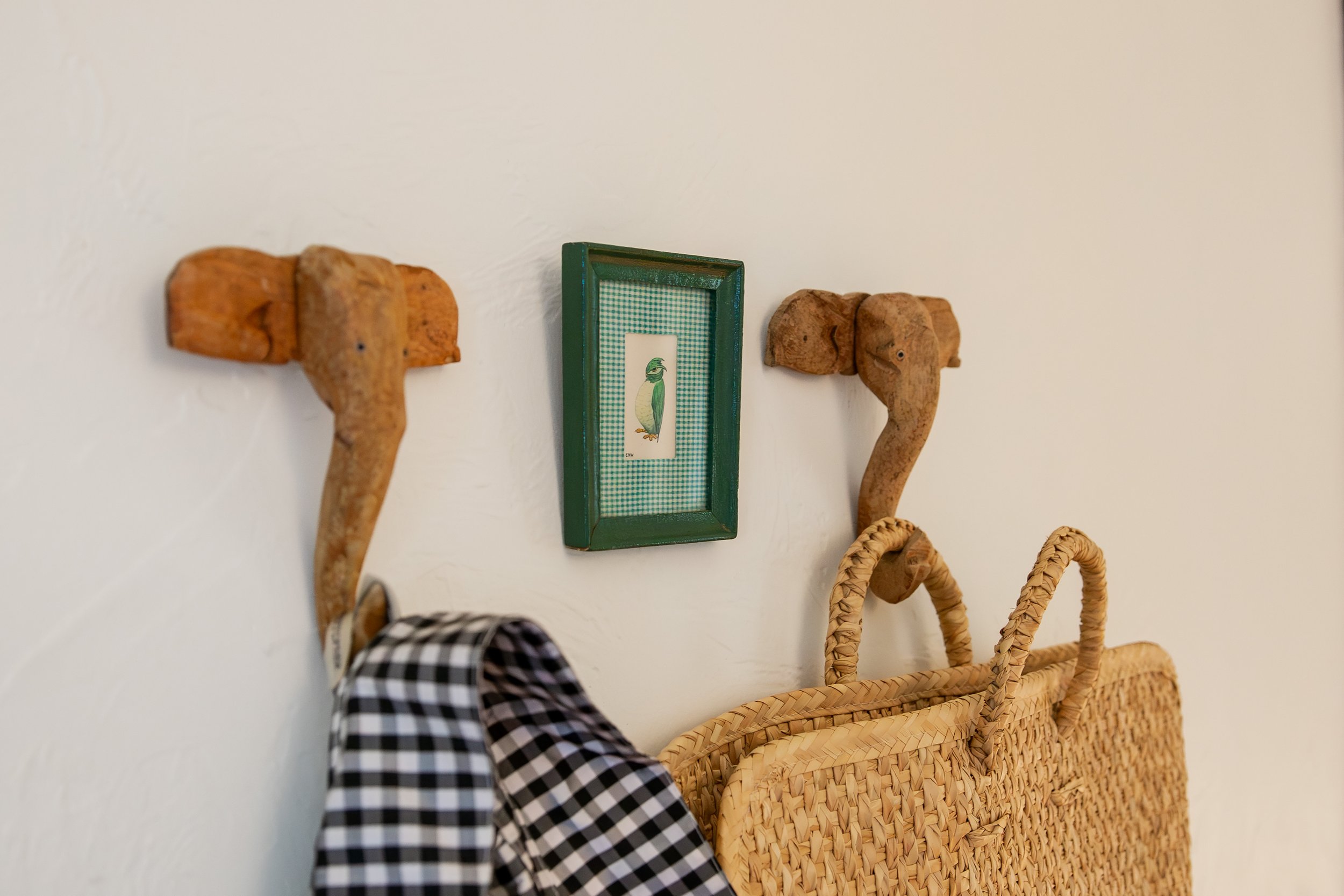 Two elephant-shaped wooden hooks on a white wall, holding a checkered cloth and a woven basket, with a small framed picture of a bird in between. Bozeman interior design, interior renovation, and interior styling. 