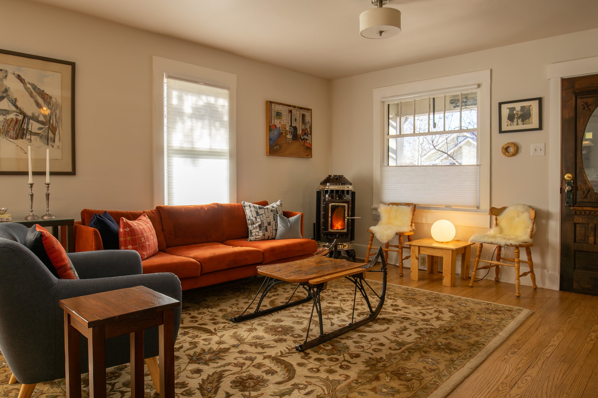 Living room with a burnt orange sofa, a gray armchair, a wooden coffee table, a rug, two chairs with sheepskin covers, and a small table with a round lamp. There is a wood stove, framed artwork, windows with blinds, and a mix of decorative items.