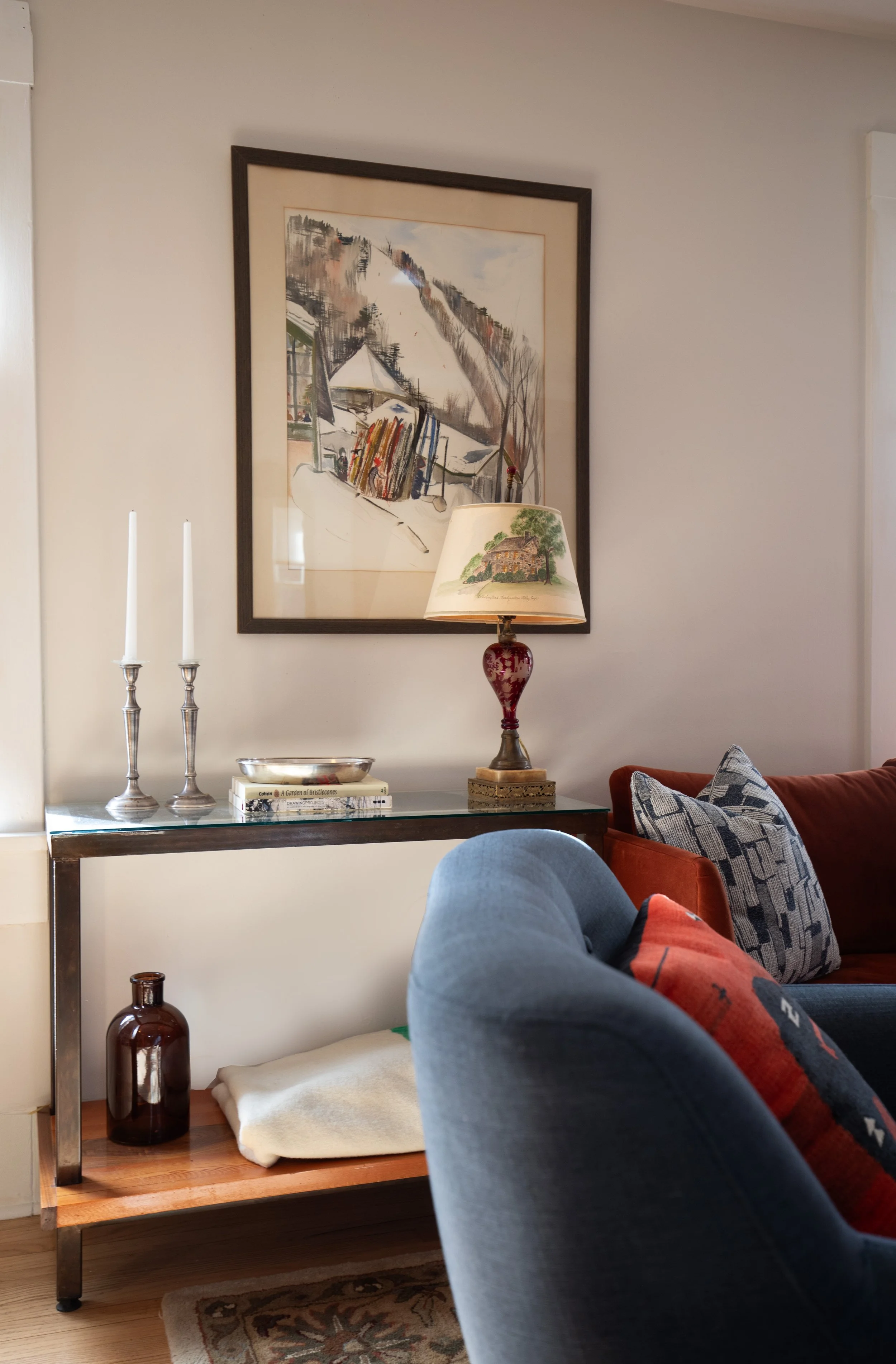 A living room corner with a glass-top side table decorated with candles, books, and a bowl. A table lamp with a traditional-style shade and a painted landscape scene is on the table. Behind the table is a large framed artwork on the wall. The corner 
