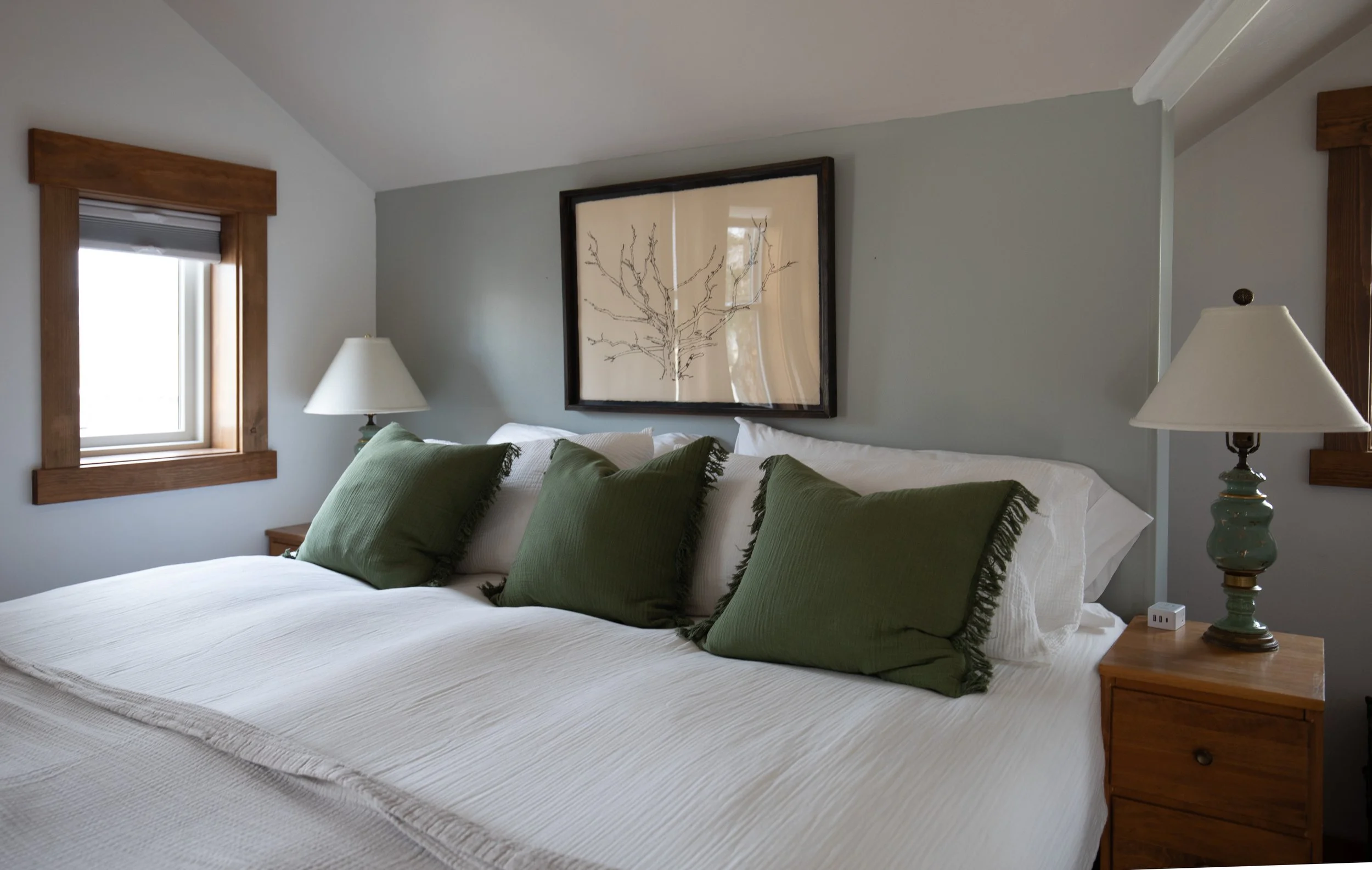 A cozy bedroom with a bed featuring white linens and three large green pillows. Wooden window frames, two beside the bed and one above, with natural light coming through. Two bedside tables with matching lamps and a framed leafless tree artwork above