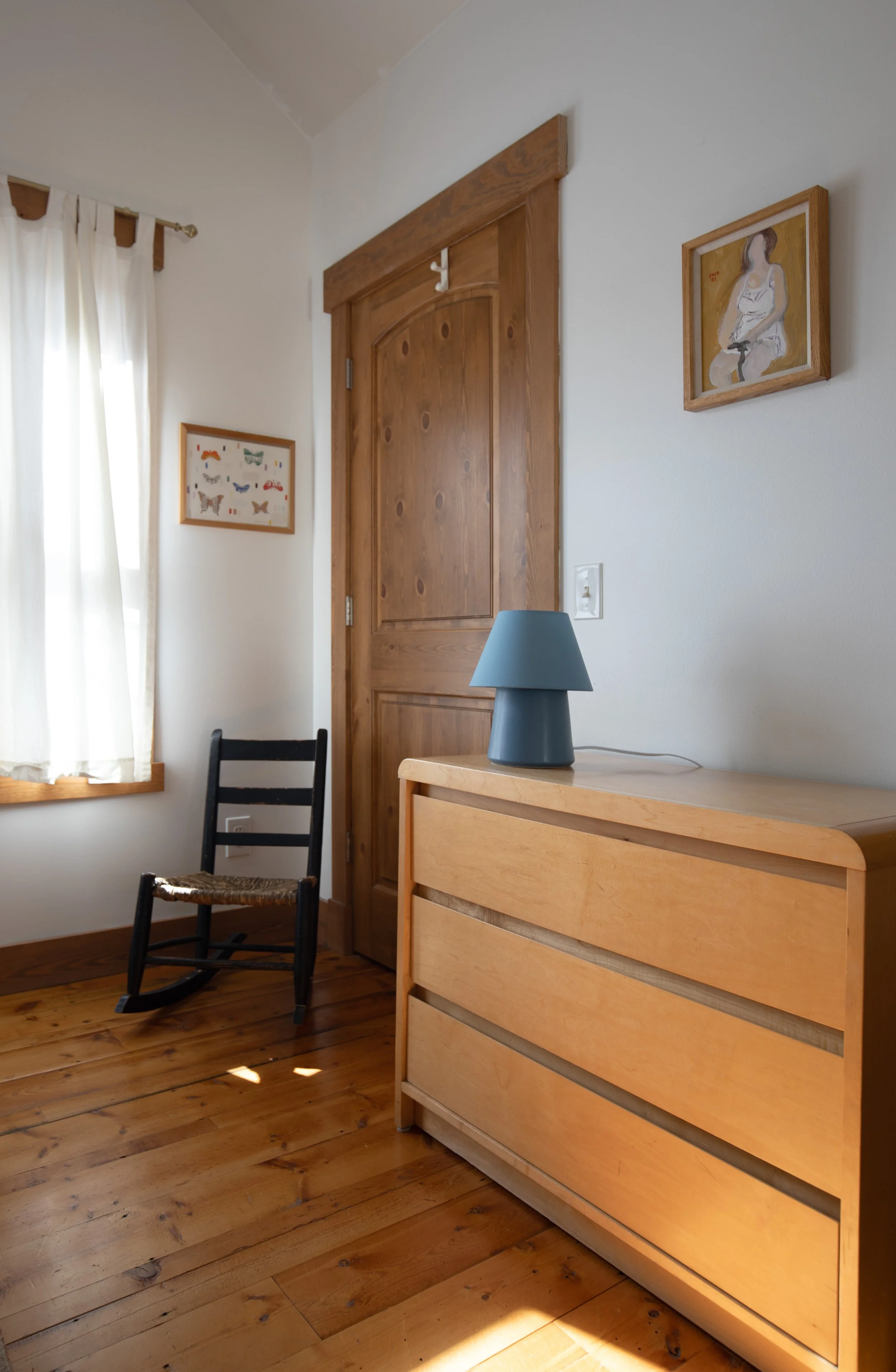 A corner of a room with a wooden floor, a wooden dresser with a blue lamp on top, a black rocking chair, a window with white curtains, and framed artwork on the wall.  refresh renovation remodel interior designer bozeman montana two dot interiors