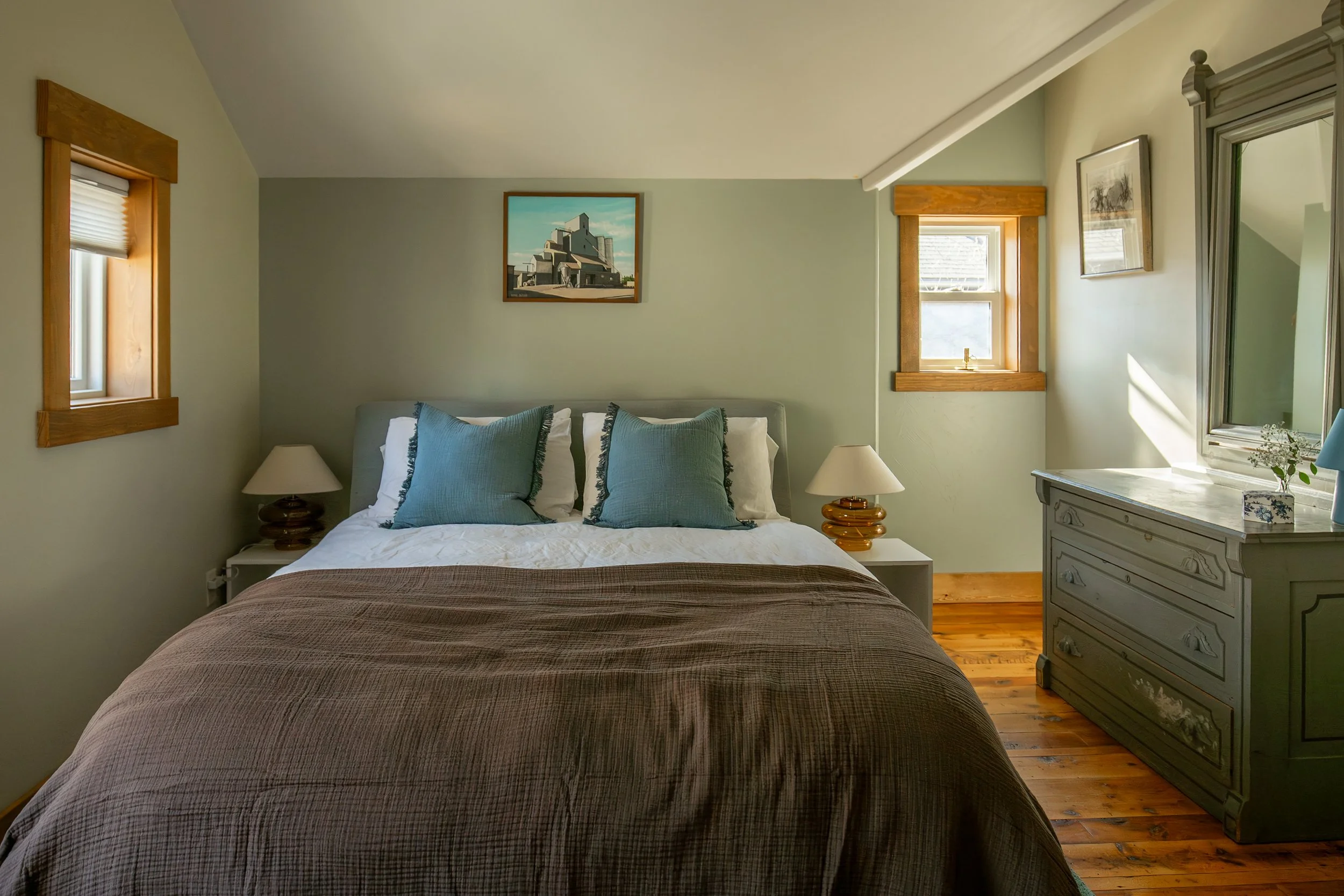 A cozy bedroom with a neatly made bed, two bedside lamps, three pillows, two windows with wooden trim, a dresser with a decorative box, and wall art, featuring a sloped ceiling and wooden flooring.  Bozeman Montana Interior Design Affordable