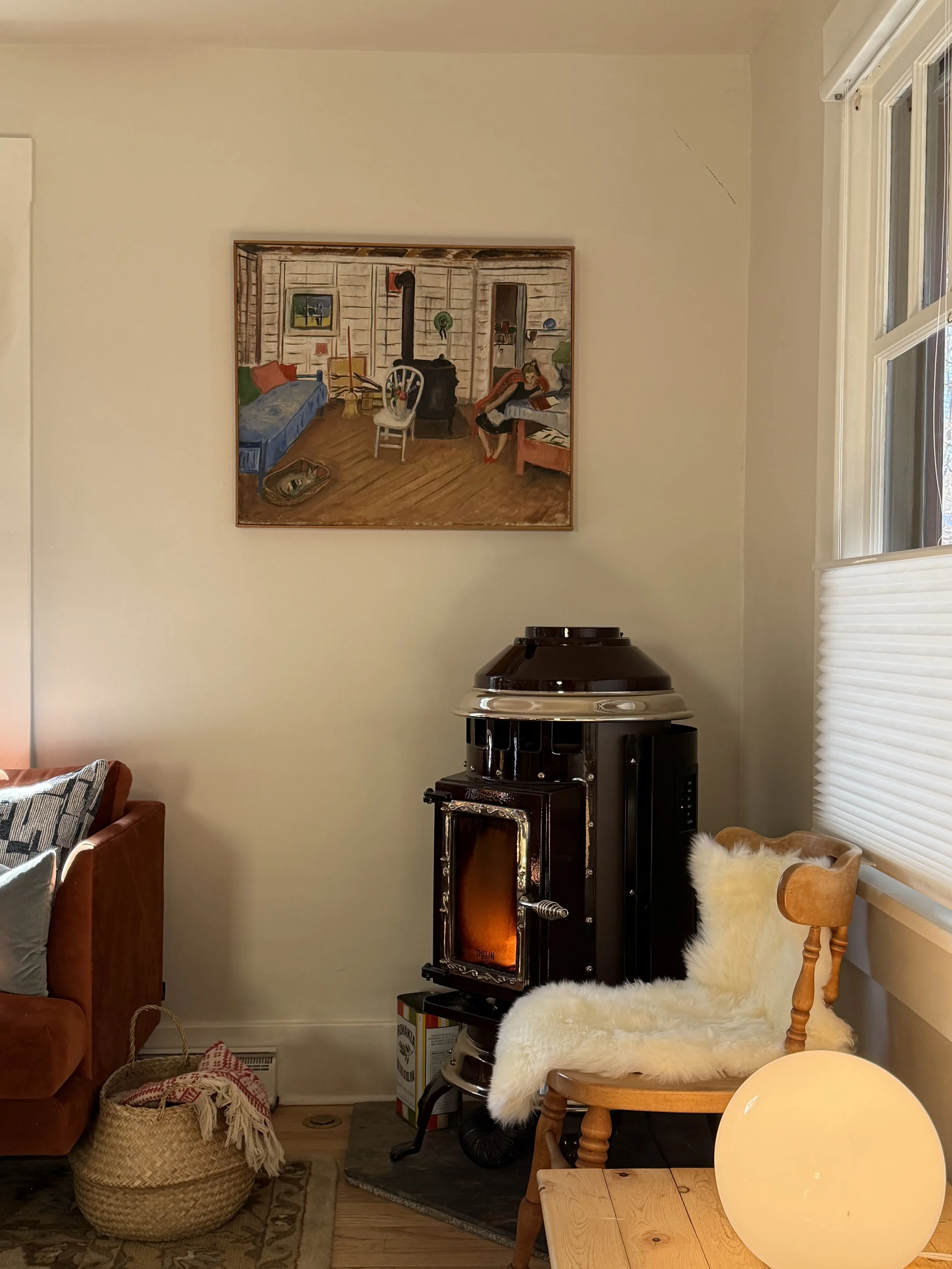 A cozy living room with a wood stove, a wooden chair with a sheepskin throw, a window with white blinds, a globe lamp, a leather sofa with patterned pillows, and a painting of an interior scene on the wall.  Bozeman Montana Interior Design Affordable