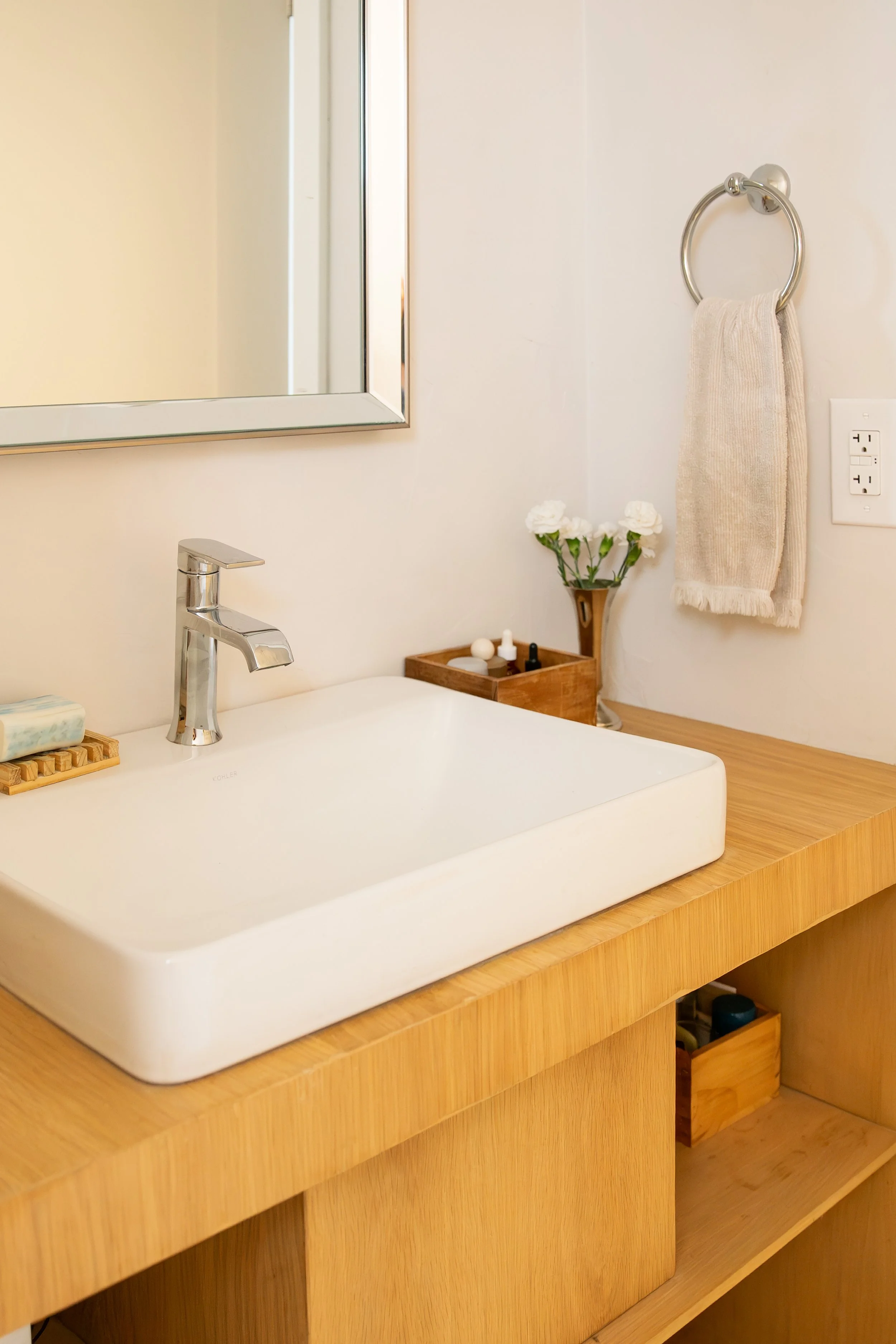 A bathroom sink with a wooden countertop, a framed mirror, a silver faucet, a small soap dish, a vase with white flowers, a wooden tray with small bottles, and a towel ring. Bozeman interior design, interior renovation, and interior styling. 