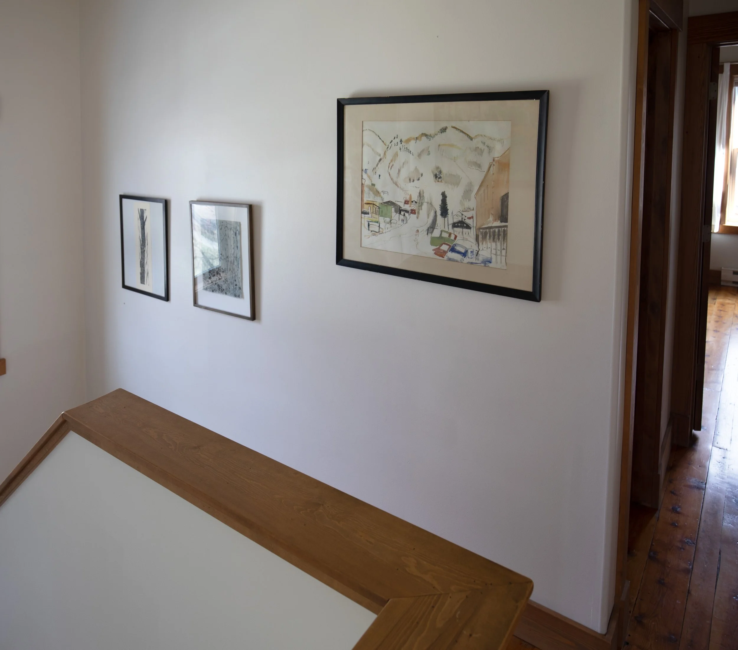 Interior hallway of a house with wooden flooring and white walls decorated with three framed artworks. A handrail of a staircase is partially visible in the foreground. STR refresh interior design bozeman montana two dot interiors