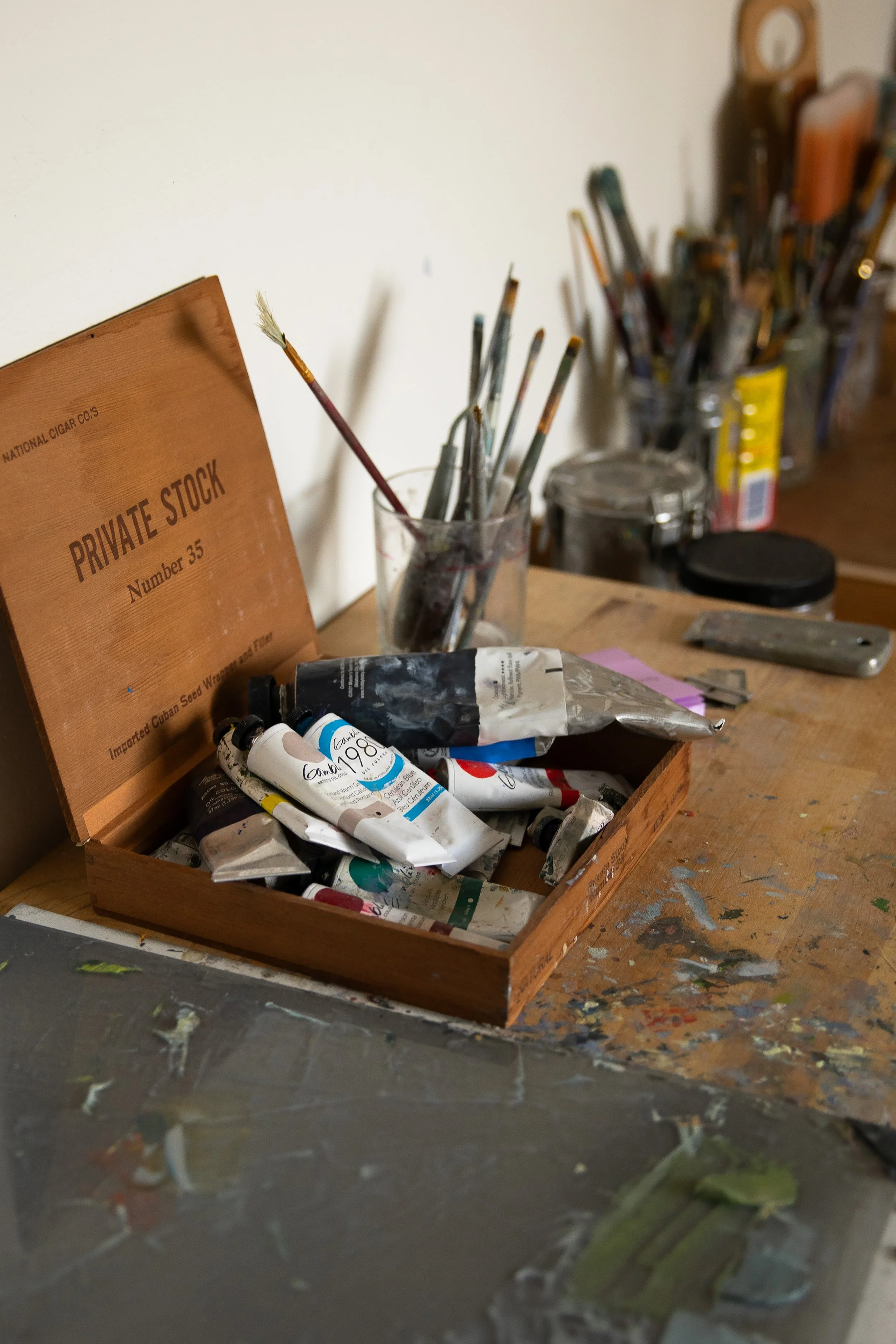 An artist's workspace with a wooden table, a box of oil paints, paintbrushes in a glass jar, and various art supplies in the background. Bozeman interior design, interior renovation, and interior styling. 