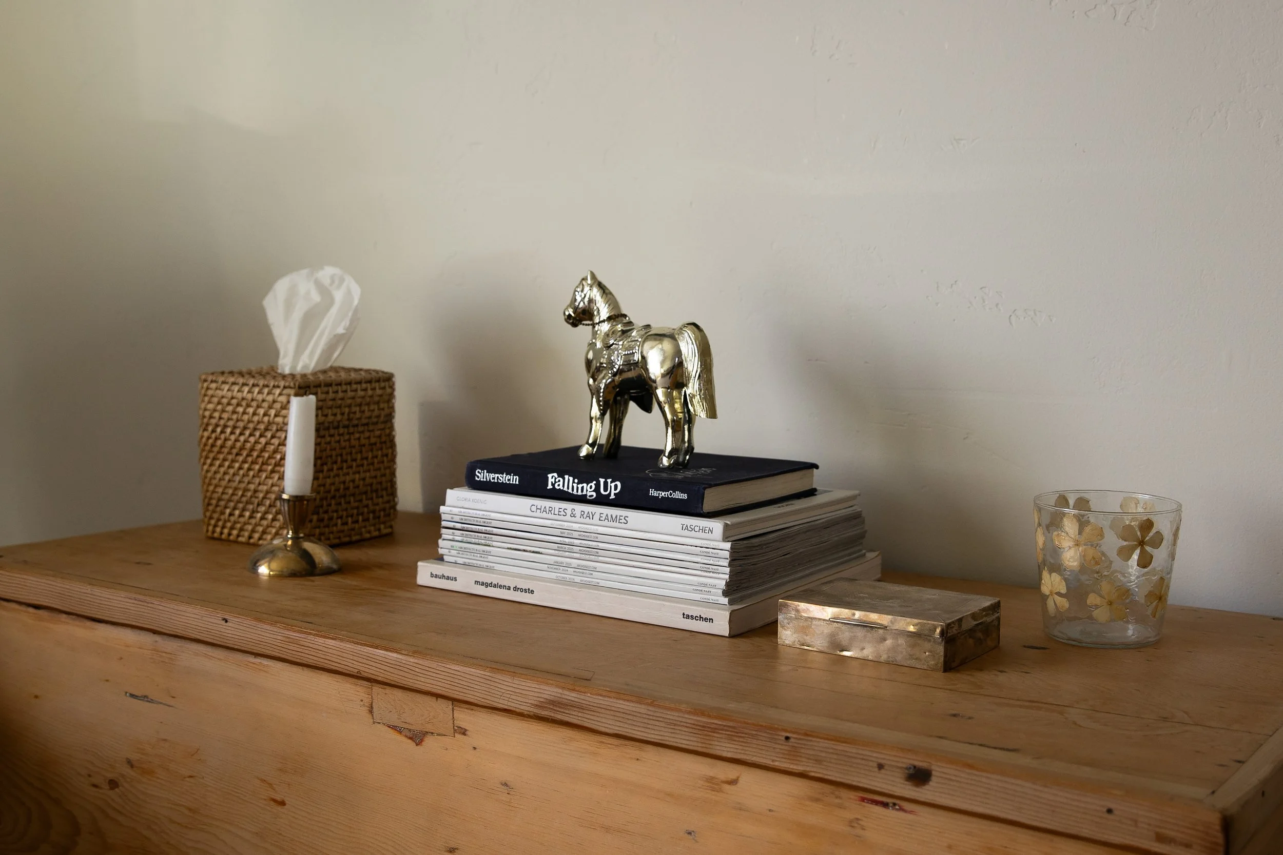 A wooden surface with a tissue box, a candle holder, a stack of books with a silver horse figurine on top, a rectangular box, and a glass with gold floral patterns. Bozeman interior design, interior renovation, and interior styling.