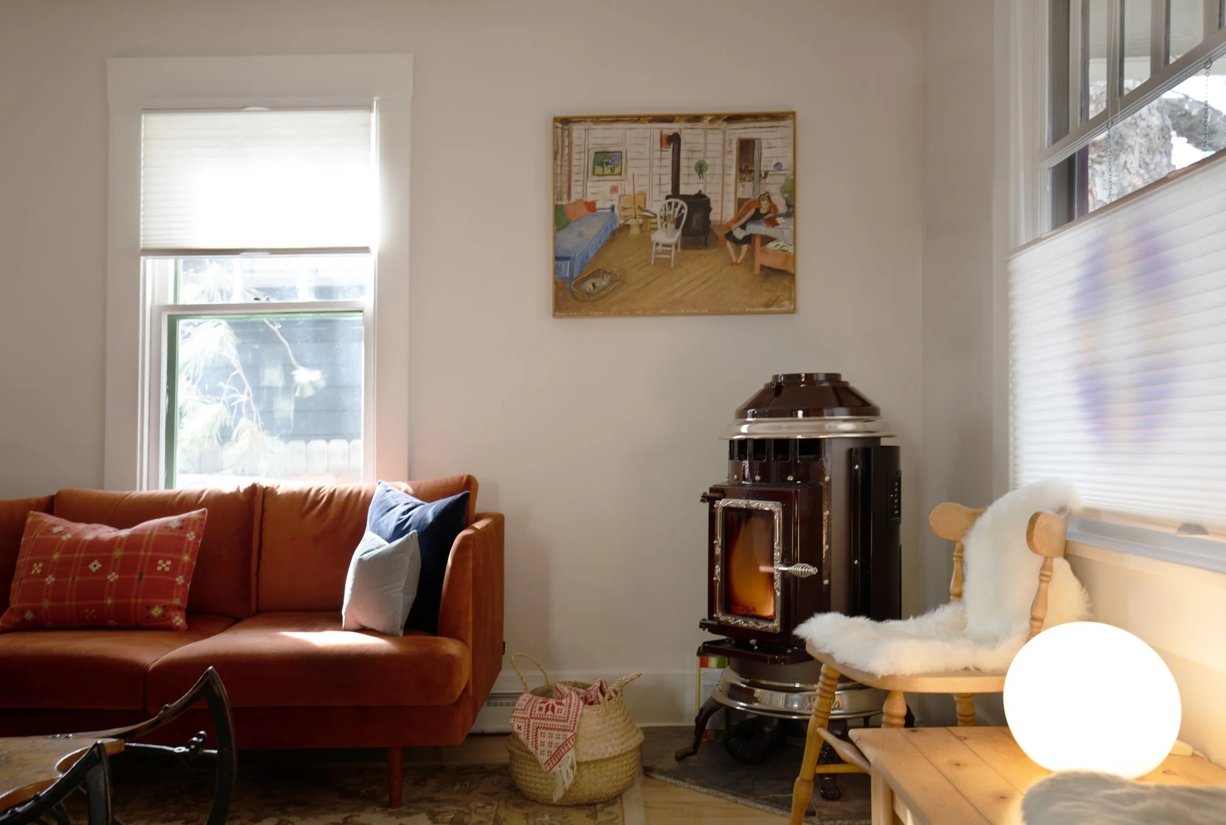 Cozy living room with a red sofa, pillows, wood chair with a sheepskin throw, a small wooden table with a globe lamp, a wood stove, framed artwork on the white wall, and windows with blinds allowing natural light. refresh renovation remodel interior 
