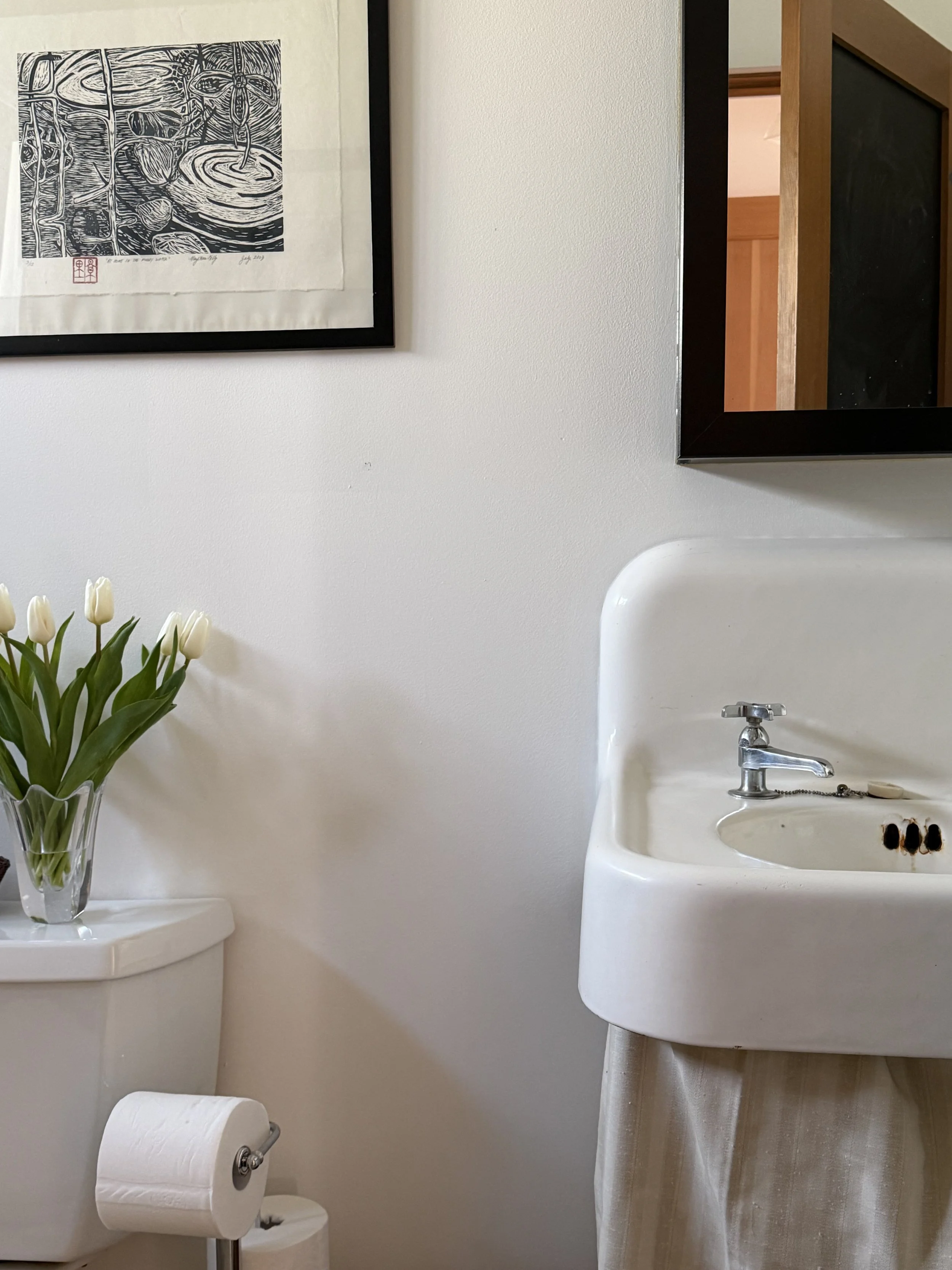 A small bathroom with white walls, a white toilet with a vase of white tulips on top, a vintage porcelain sink with a silver faucet, and a mirror above the sink. There is a framed black-and-white artwork hanging on the wall.