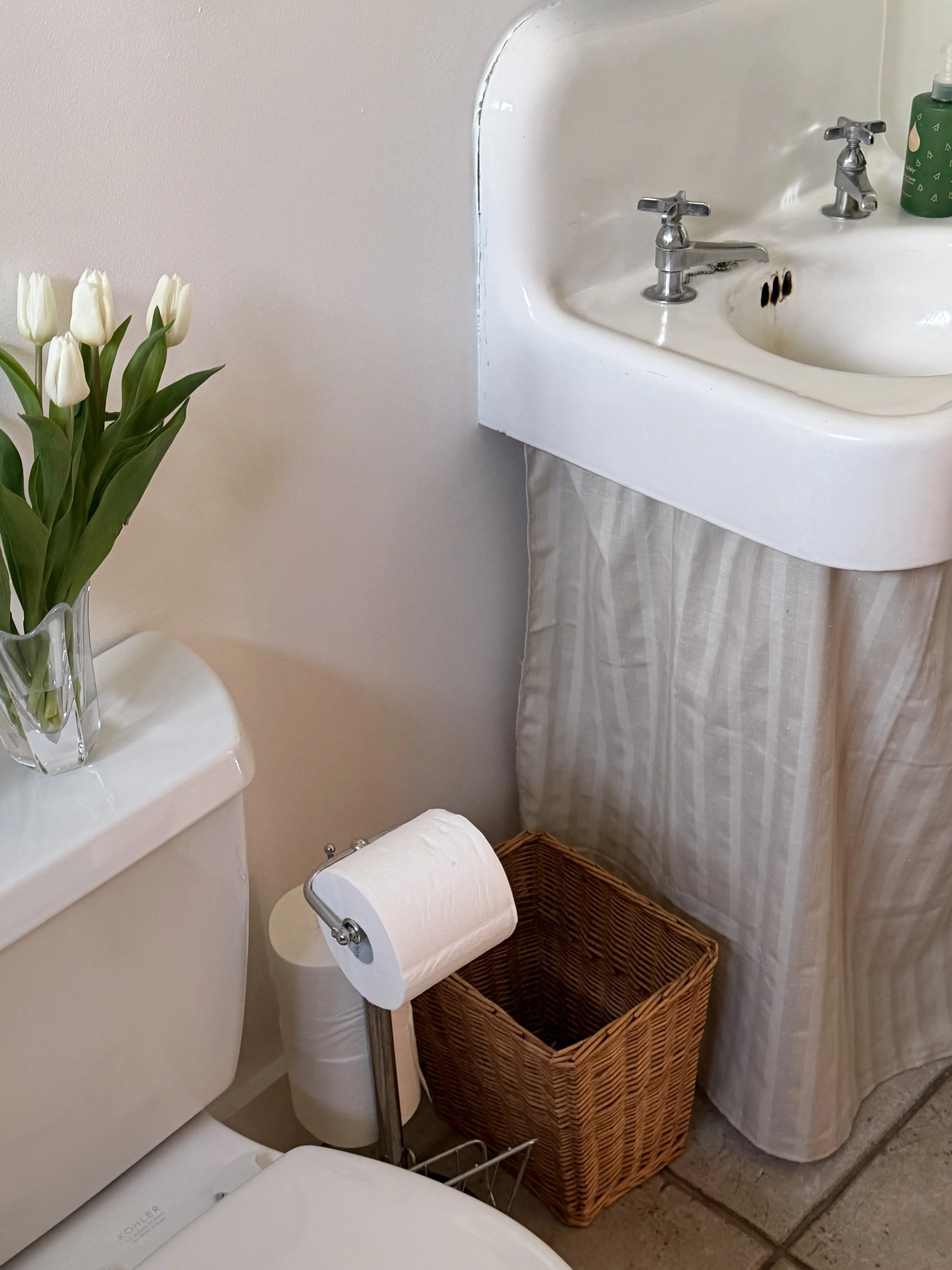 A small bathroom , vintage white porcelain sink with chrome taps, a wicker trash bin, a toilet with a roll of toilet paper on a stand, and a vase of white tulips. A beige curtain covers the lower part of the sink cabinet. Bozeman Montana Affordable