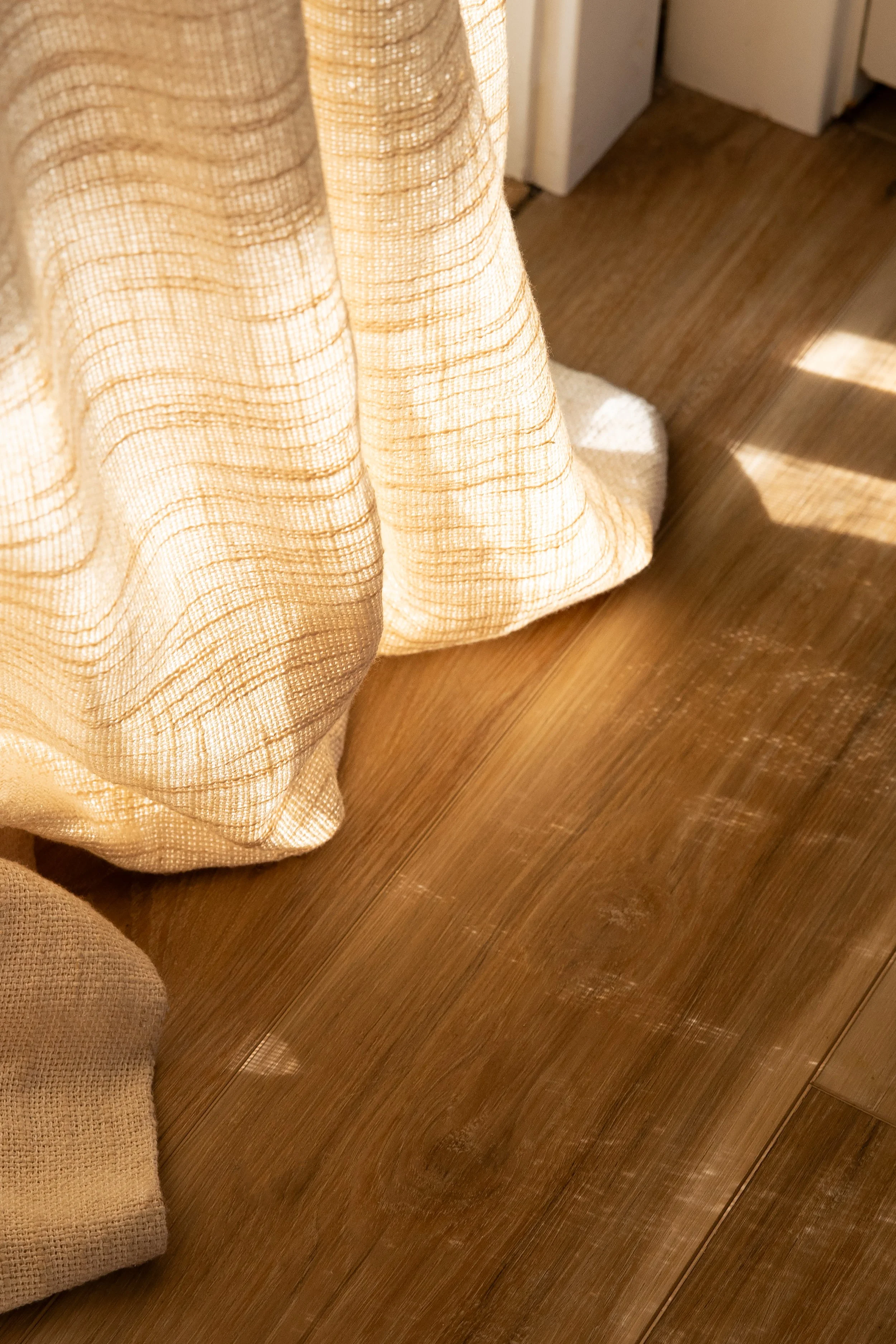 A beige curtain hanging from the ceiling, partially covering a window, with sunlight creating shadows on a wooden floor. Bozeman interior design, interior renovation, and interior styling. 