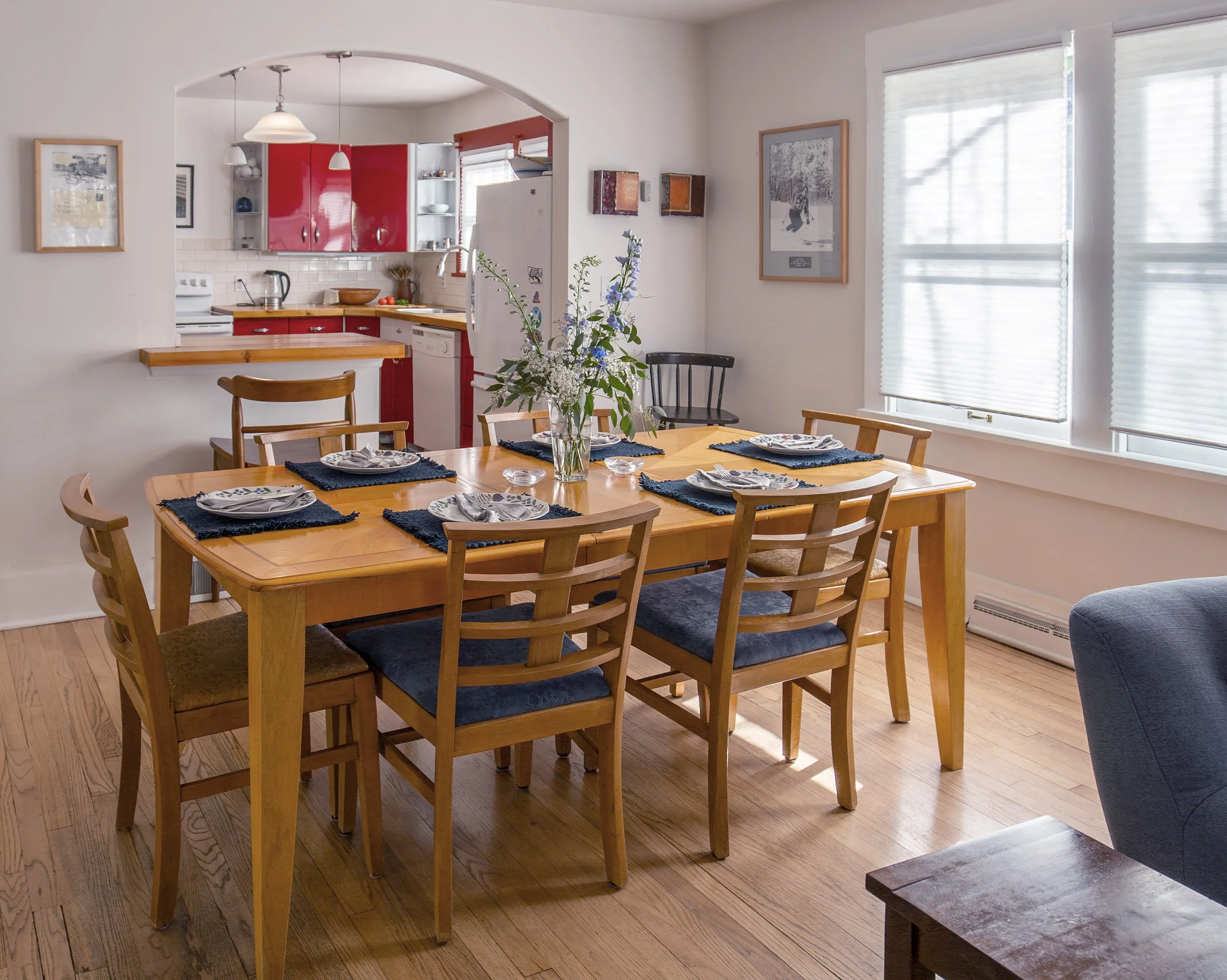 A dining room with a wooden table set for six, featuring plates, cutlery, and blue placemats, with a vase of flowers in the center. The room has large windows with blinds, and an open kitchen with red cabinets is visible in the background. refresh