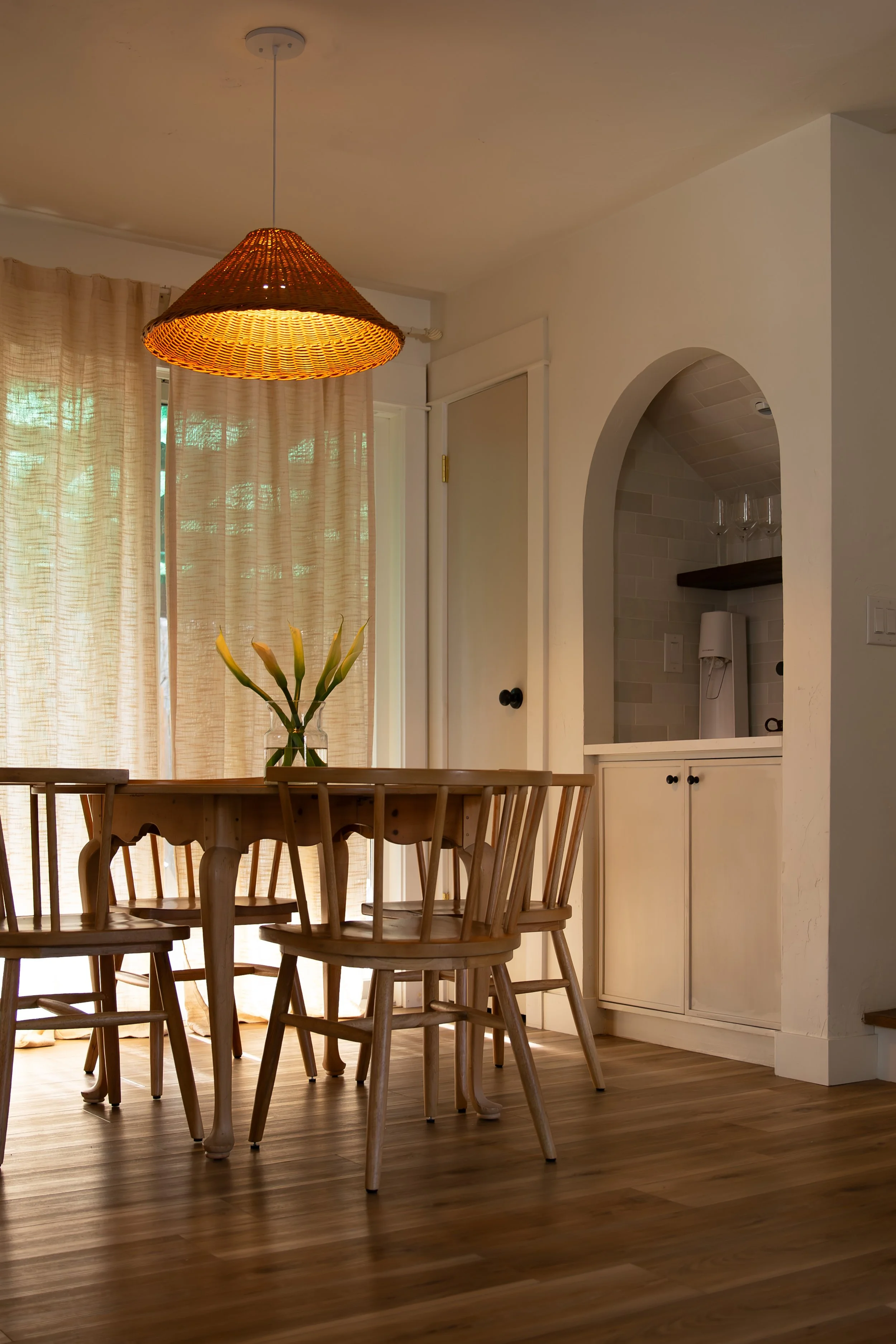 Dining room with wooden table and chairs, vase with calla lilies, beige curtains, and a hanging woven lamp. Bozeman interior design, interior renovation, and interior styling. 