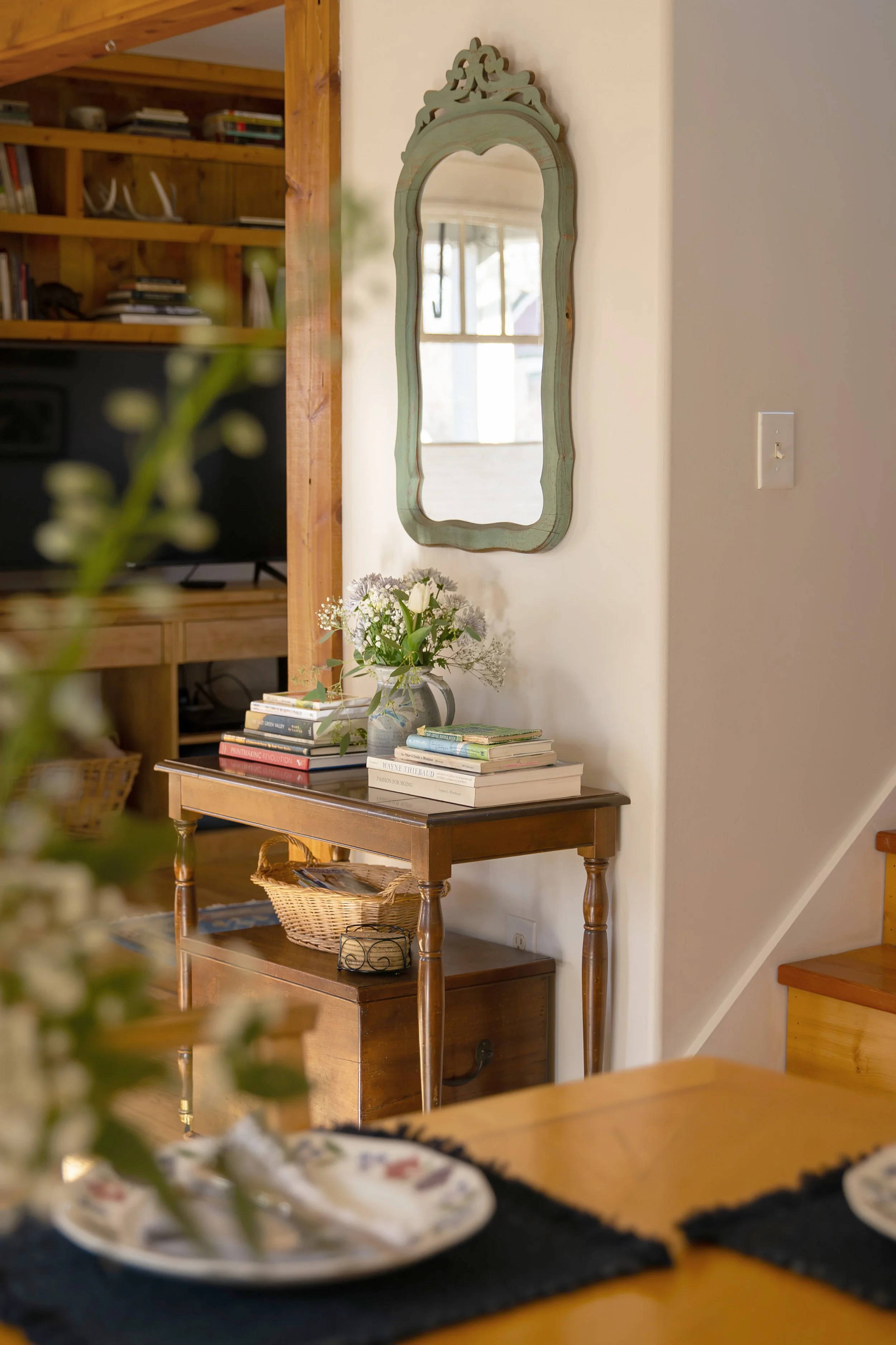 Interior of a cozy home with a wooden console table holding a vase of white flowers and stacked books. A decorative mirror hangs above the table, reflecting a window. Part of a staircase is visible in the background.