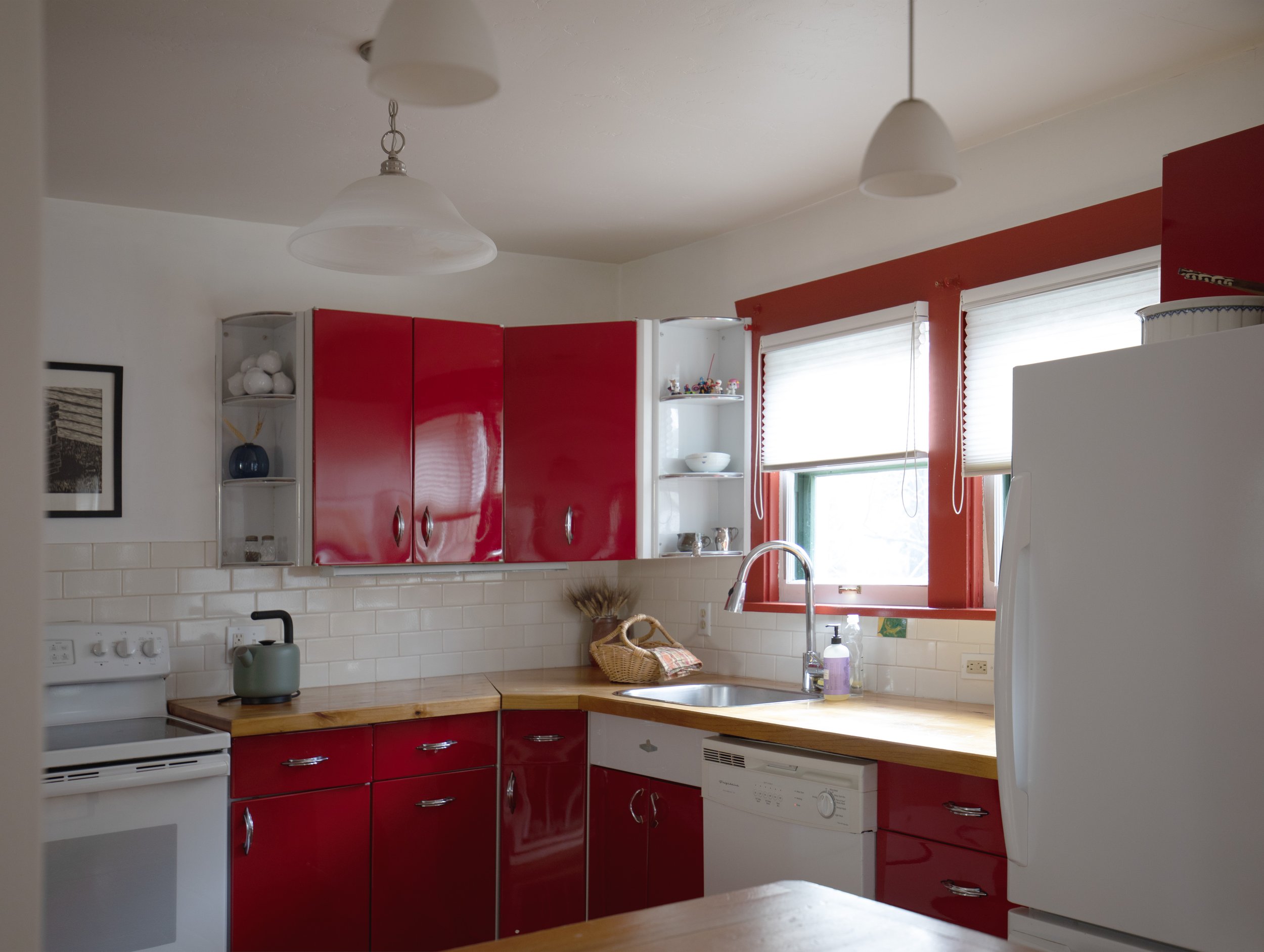 Kitchen with white walls, red cabinets, and wooden countertops. There's a white stove, a microwave, a dishwasher, a white refrigerator, and two windows with white blinds. Decor includes a framed picture, a basket, and small decorations on open shelve
