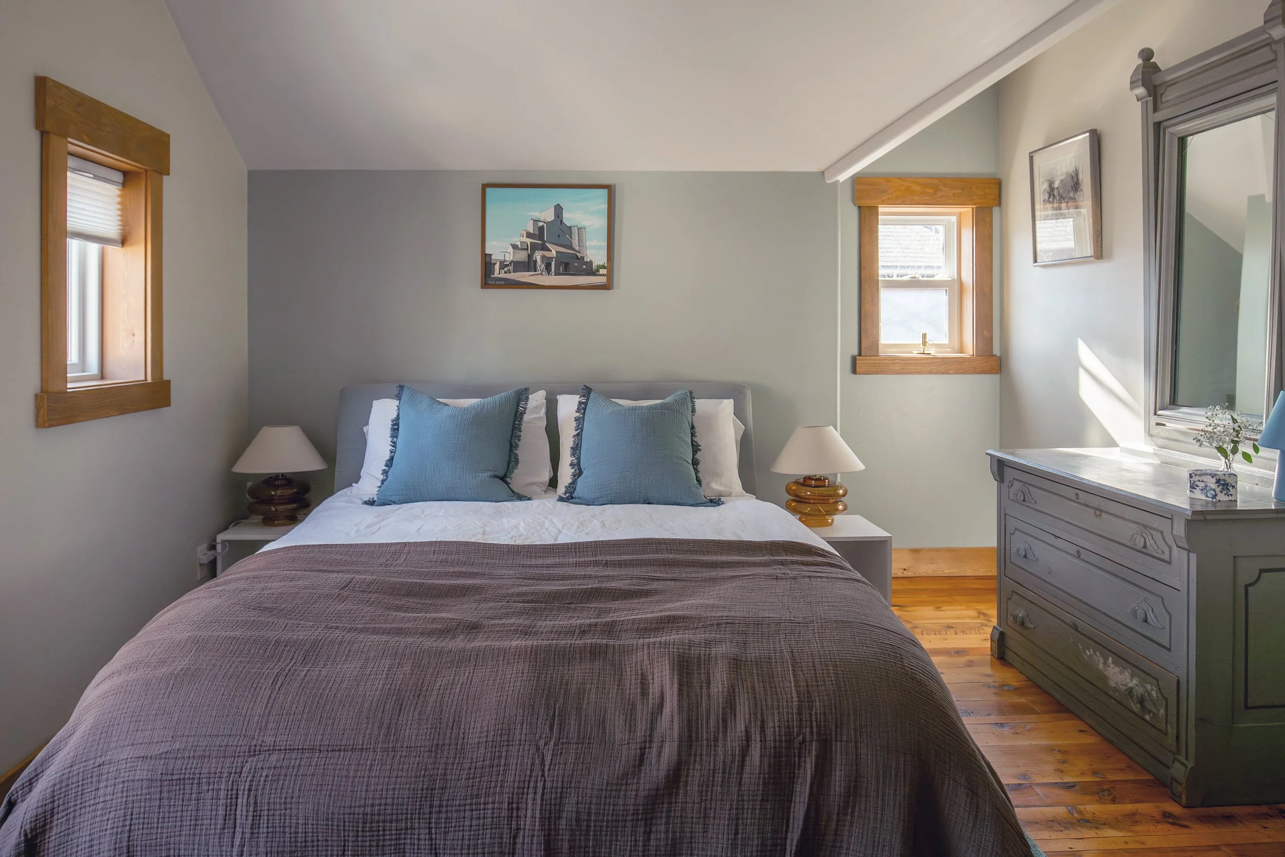 A cozy bedroom with a bed, blue pillows, white bedsheets, wooden nightstands, lamps, a dresser, and windows with wooden frames. refresh renovation remodel interior designer bozeman montana two dot interiors