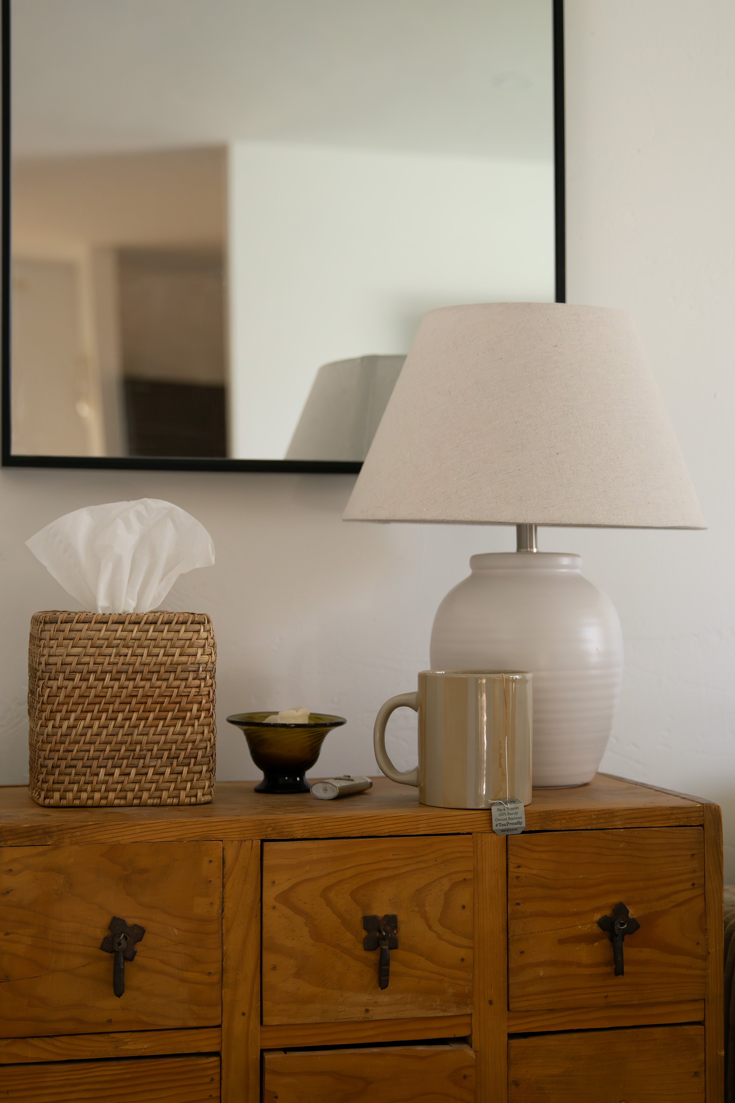 A wooden dresser with a beige lamp, a coffee mug, a small black dish, a tissue box, and small objects, with a mirror reflecting a light-colored wall behind. Bozeman interior design, interior renovation, and interior styling. 