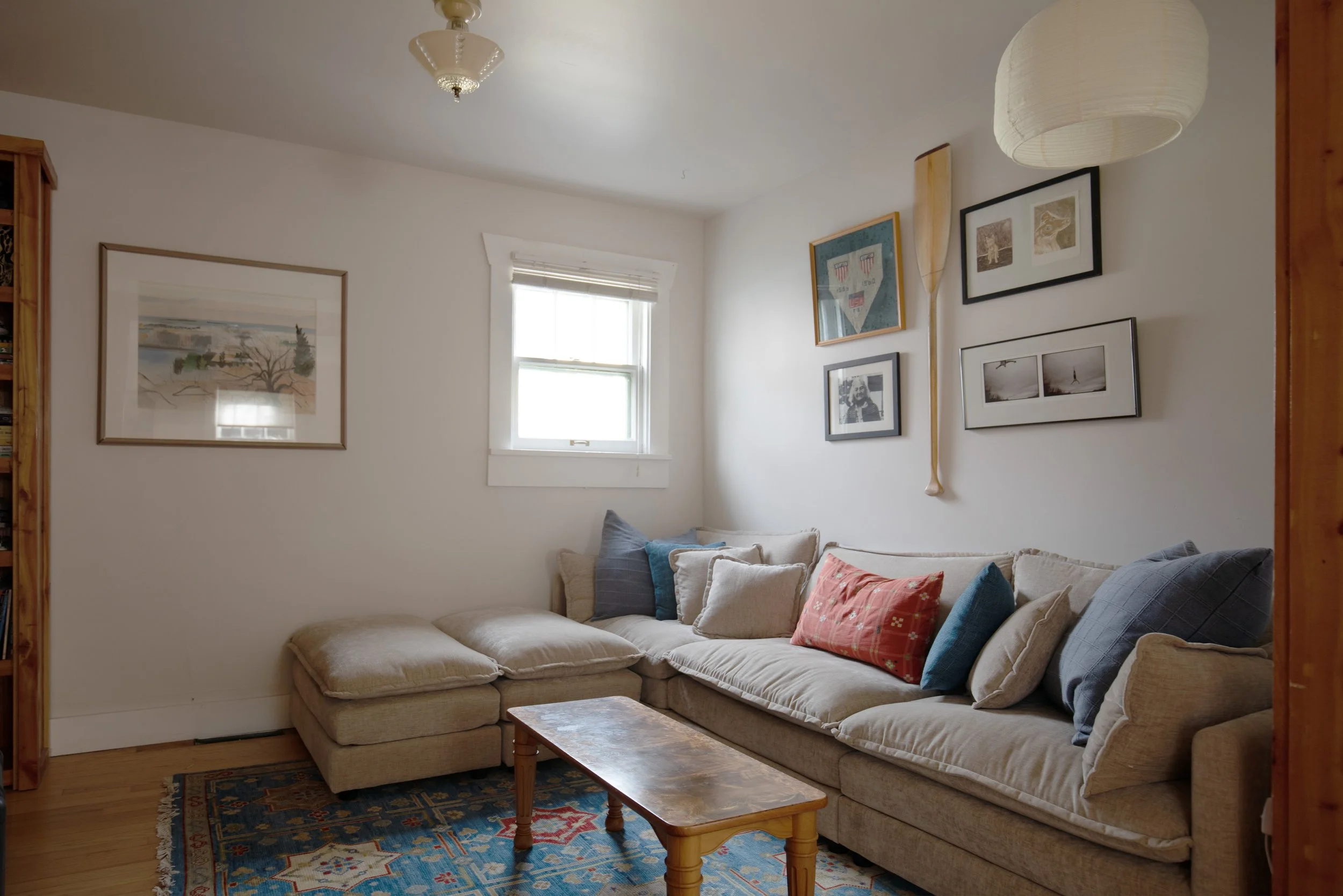Living room with beige L-shaped sofa adorned with multiple pillows, a wooden coffee table, a patterned area rug, a window with white trim, and wall art including framed pictures and a decorative paddle.