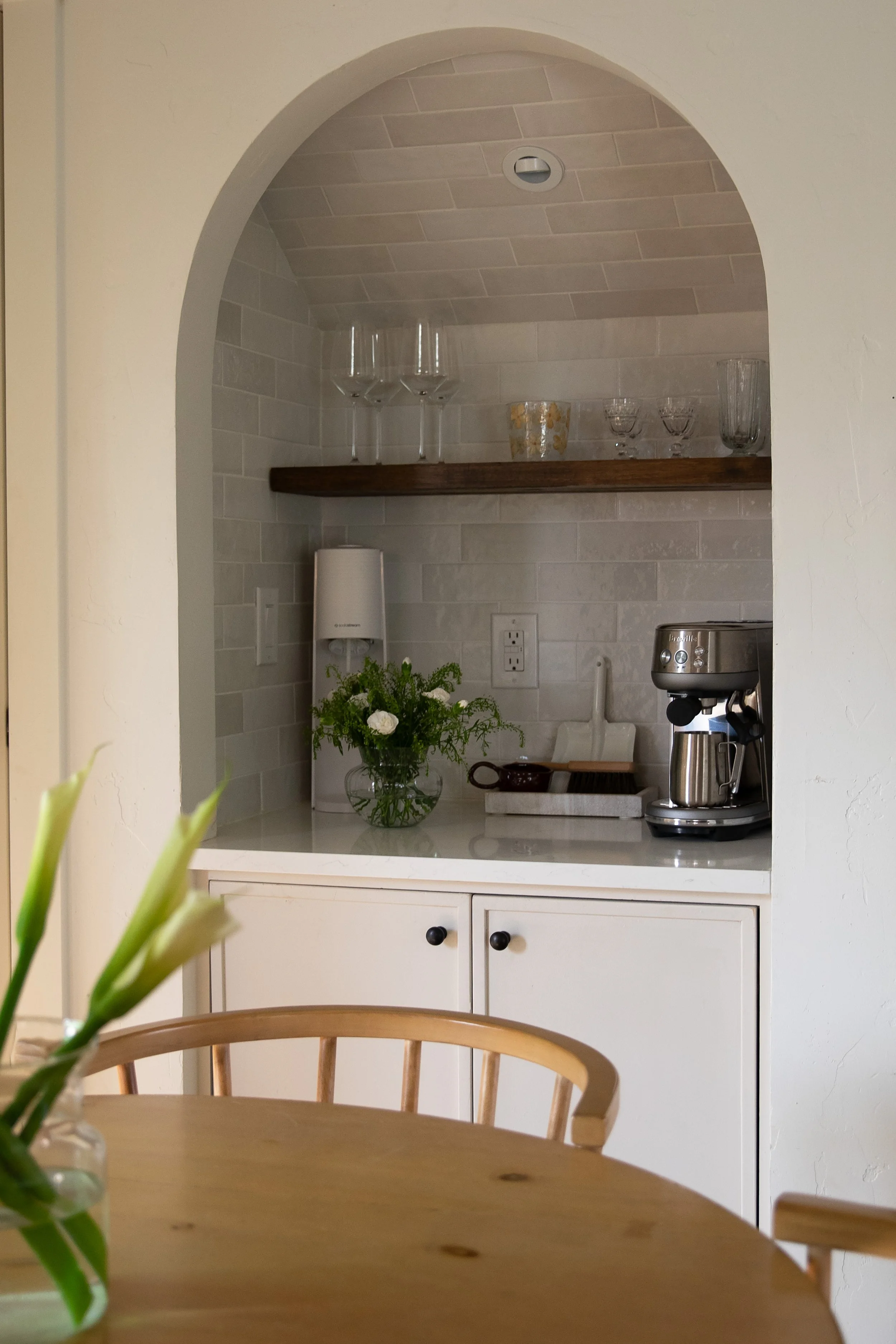 A kitchen nook with a white cabinet and countertop, a coffee maker, a vase with white flowers, and utensils. A wooden shelf above holds glassware and decorative items. Bozeman interior design, interior renovation, and interior styling. 