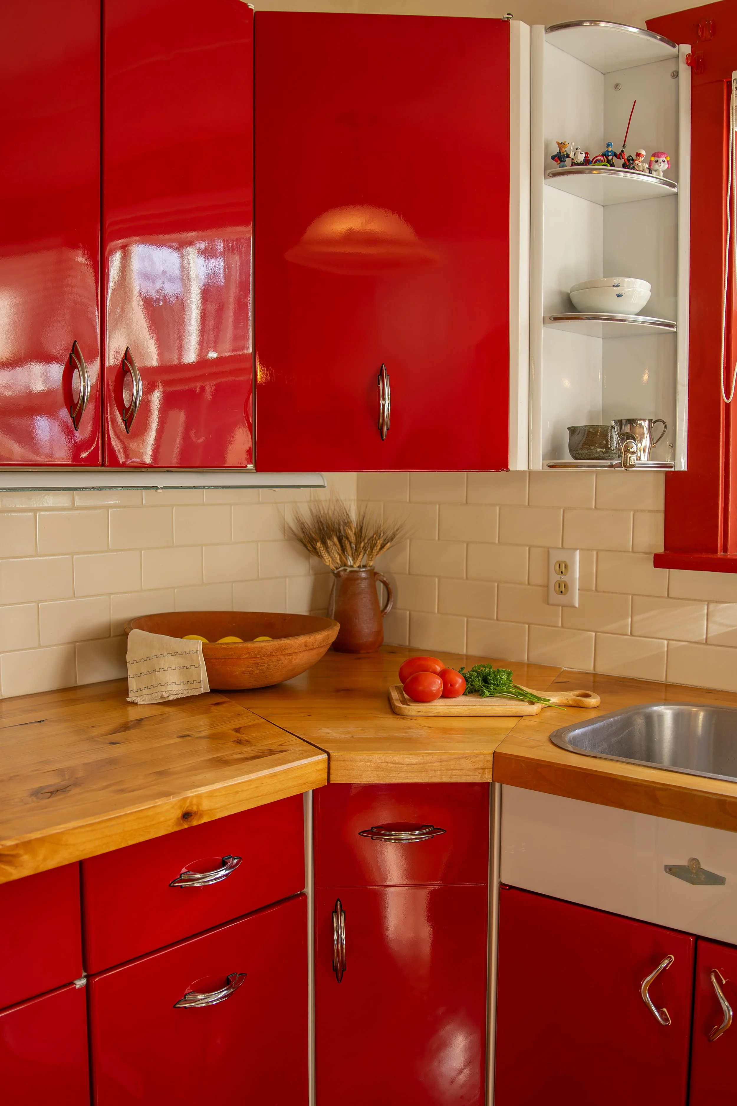 A kitchen corner with red cabinets, wooden countertop, a sink, and a small open shelf with toys, dishes, and a cup. Fresh tomatoes and parsley are on a cutting board.