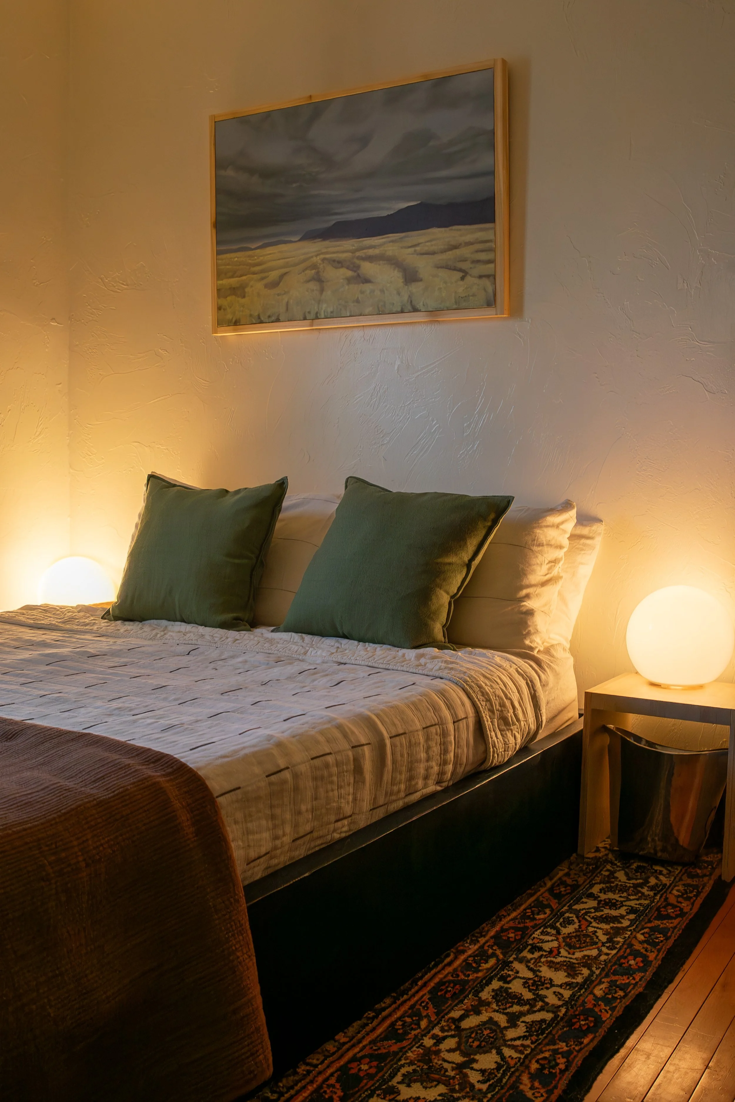 A cozy bedroom with a bed featuring green pillows and a beige quilt, illuminated by two round bedside lamps, with a painting of a landscape hanging above the bed. Bozeman interior design, interior renovation, and interior styling. 