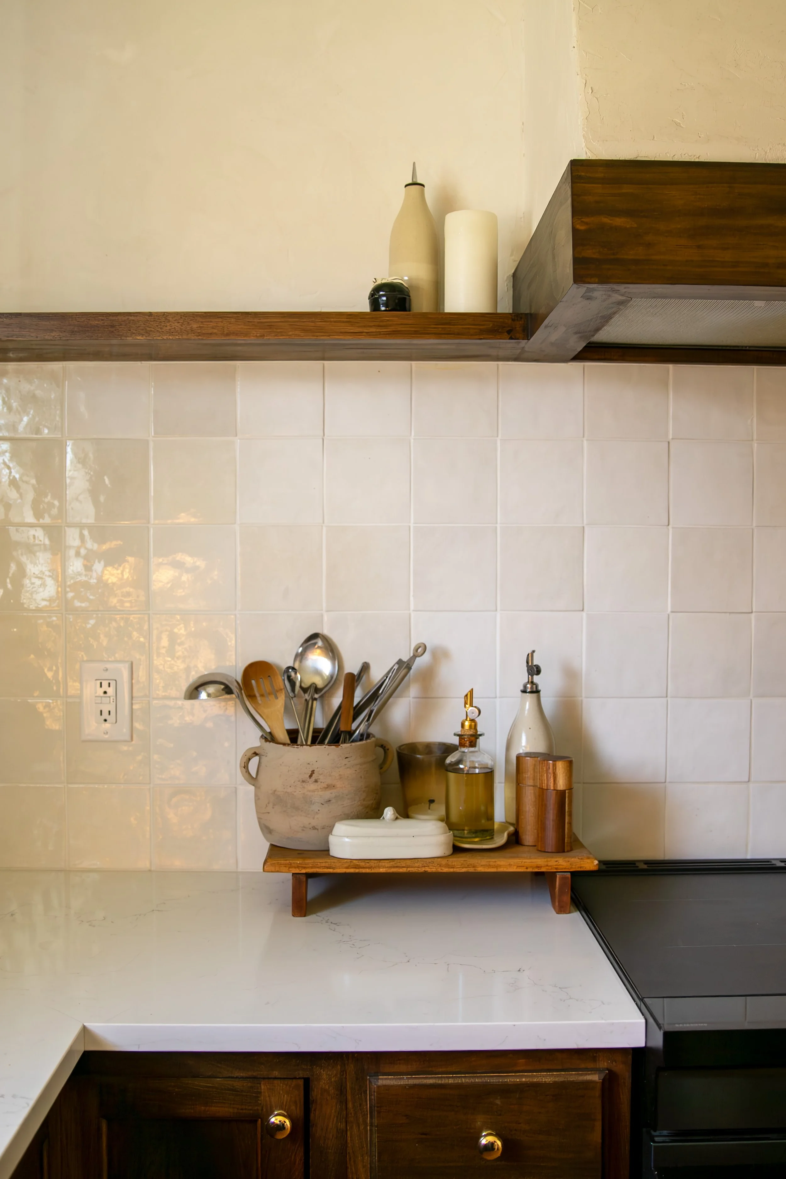 Kitchen countertop with utensils in a pottery jar, a small wooden shelf holding oil, beige tiled wall, and part of a stove. Bozeman interior design, interior renovation, and interior styling. 