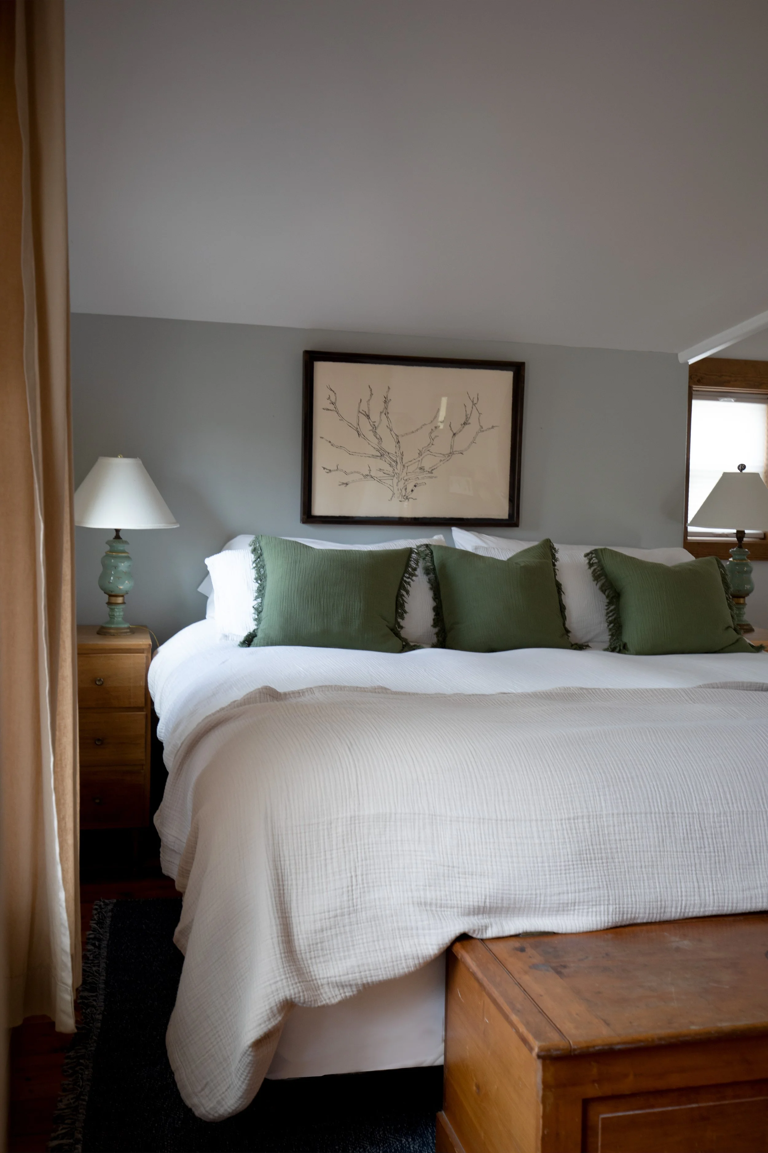 A cozy bedroom with a neatly made bed, four green pillows, white bedding, a framed artwork of a tree branch, and two matching lamps on wooden nightstands. refresh renovation remodel interior designer bozeman montana two dot interiors