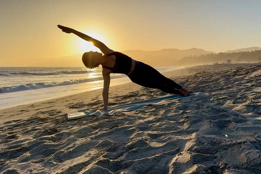 Person practicing yoga on the beach during sunset, performing a side plank pose with an ocean and mountain background.