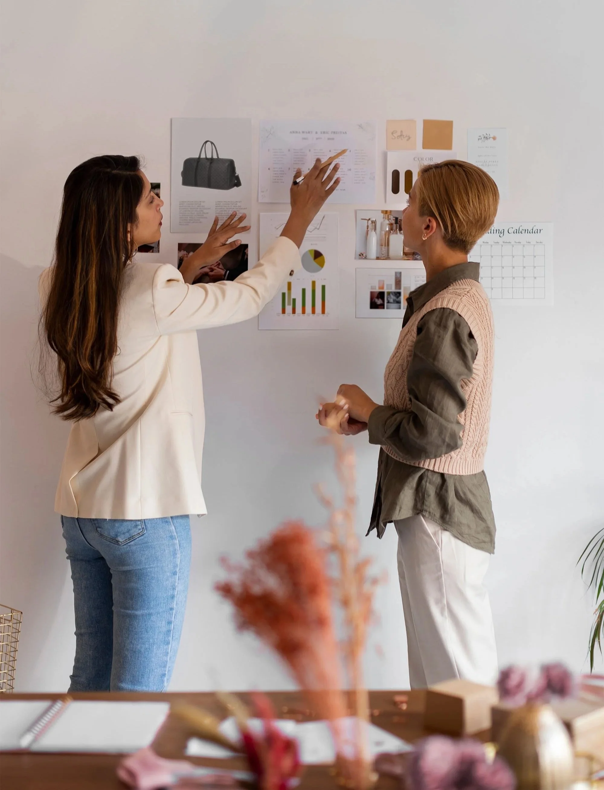 Two women in a discussion in front of a white wall with various pinned images and documents, engaged in a conversation or presentation.
