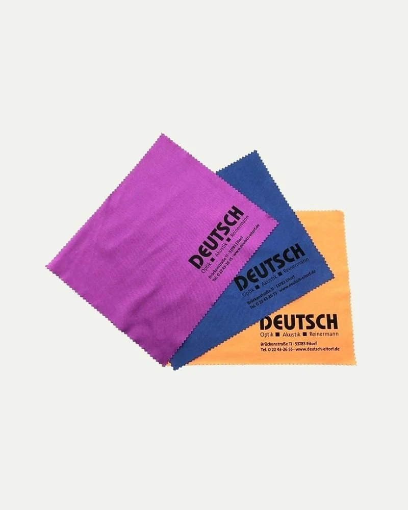 Three colorful cleaning cloths in purple, blue, and orange, with German text and a company logo for an optician and hearing specialist.